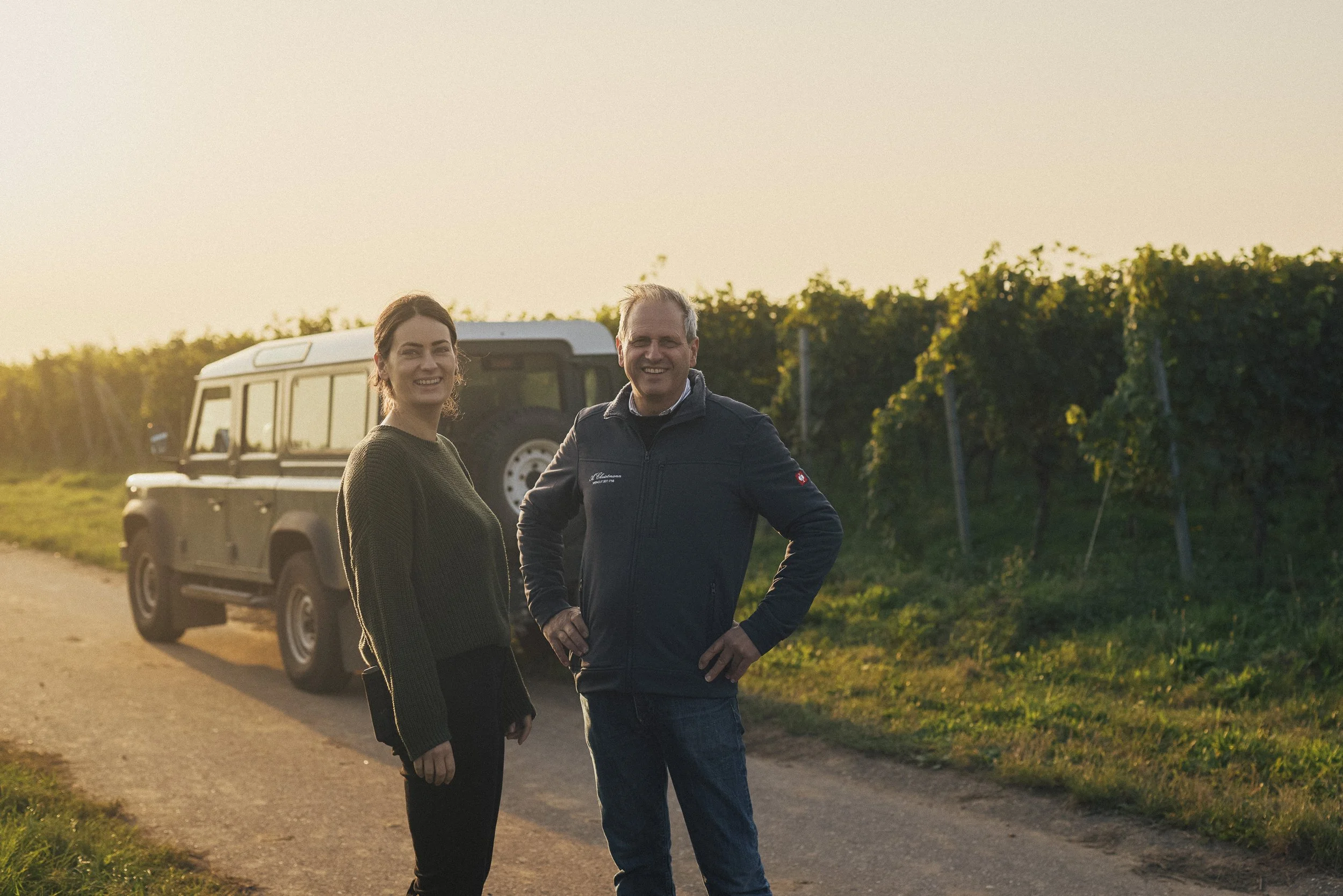 Zwei Menschen stehen neben einem Auto auf einer ländlichen Straße bei Sonnenuntergang, umgeben von Weinreben.