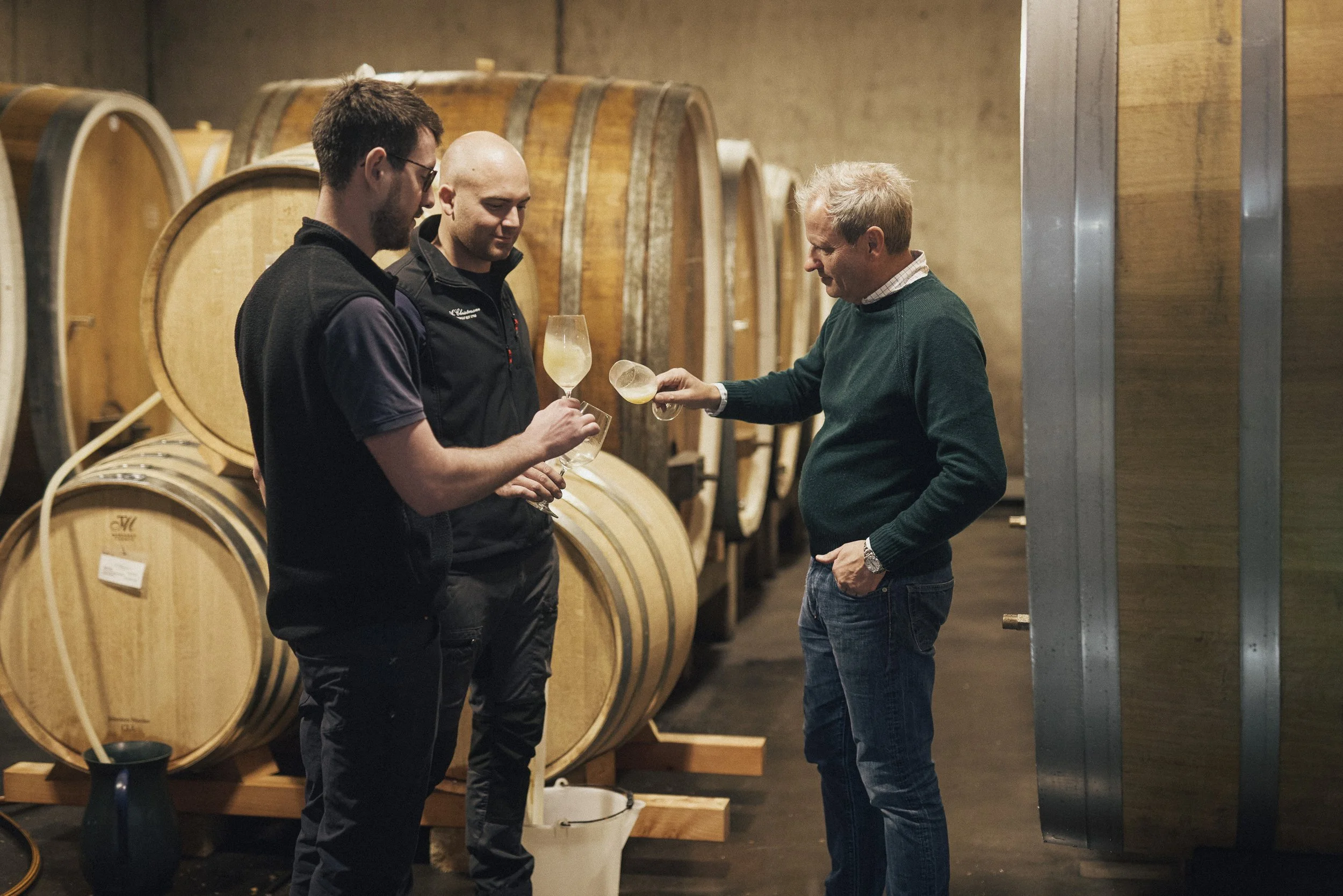 Drei Männer in einer Weinkellerei, die sich Weinproben teilen, mit Weinfässern im Hintergrund.