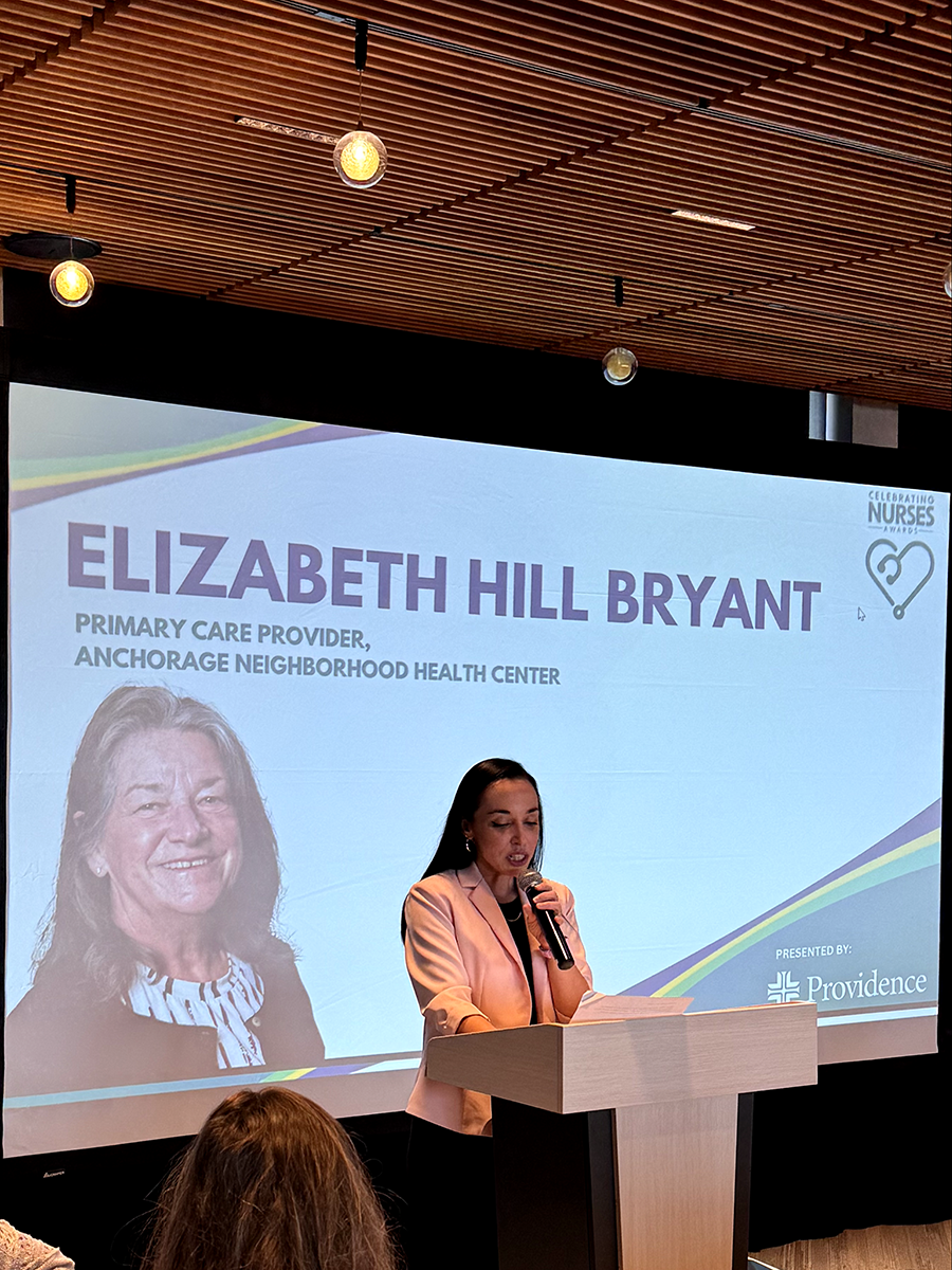 A presenter speaks at the podium as Elizabeth Hill-Bryant, FNP-C, is recognized on screen during the Celebrating Nurses Awards event.