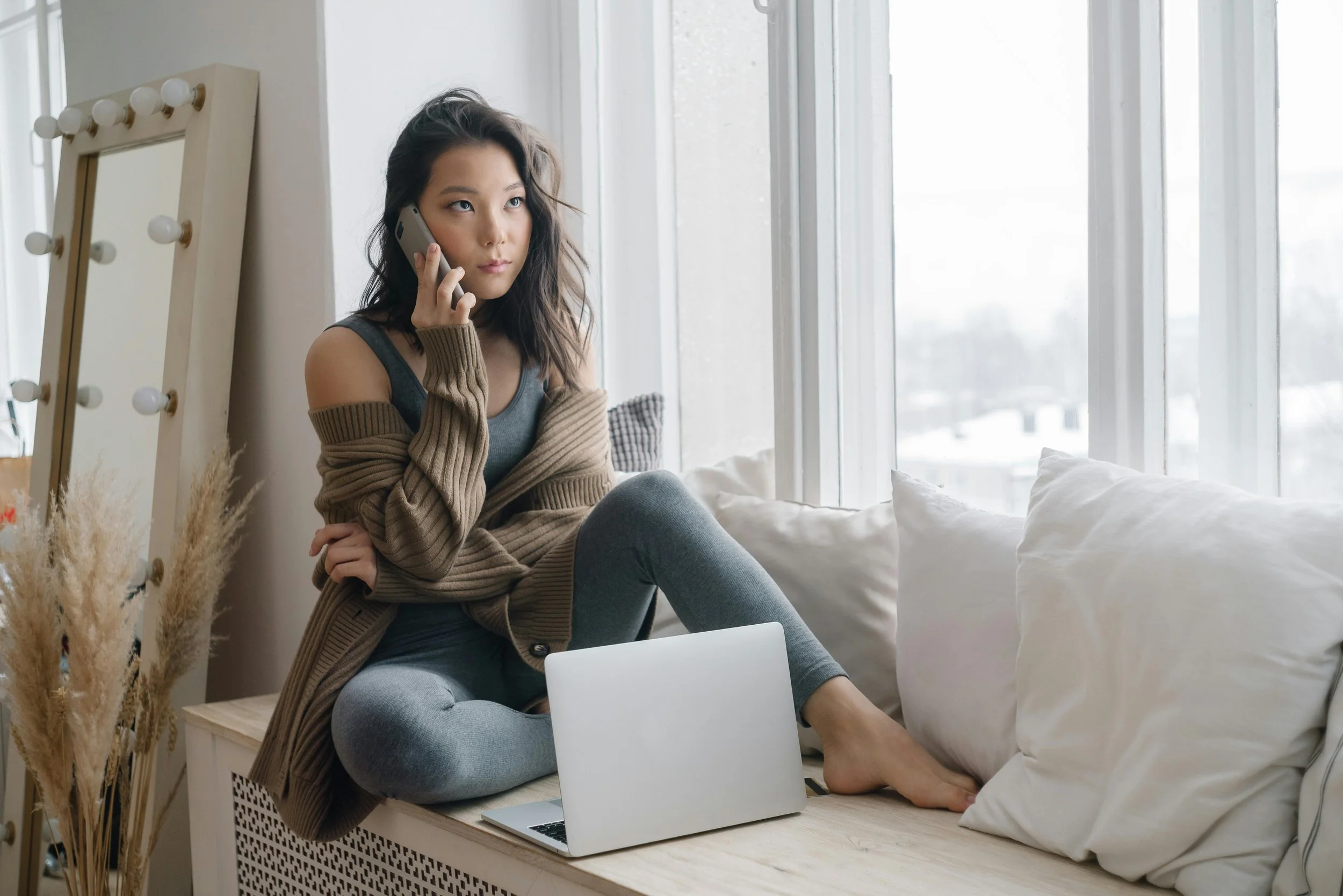 Woman sitting by window on phone with laptop, representing virtual therapy and seeking support for anxiety and relationship stress