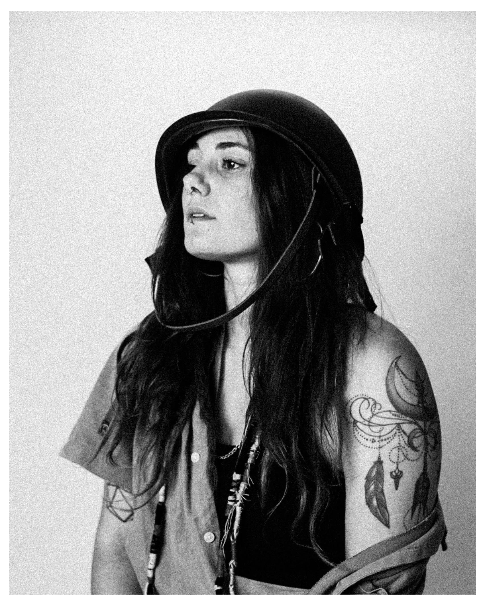 Black and white photo of a woman with long dark hair wearing a helmet, with a tattoo sleeve on her right arm featuring feathers and a crescent moon, against a plain background.