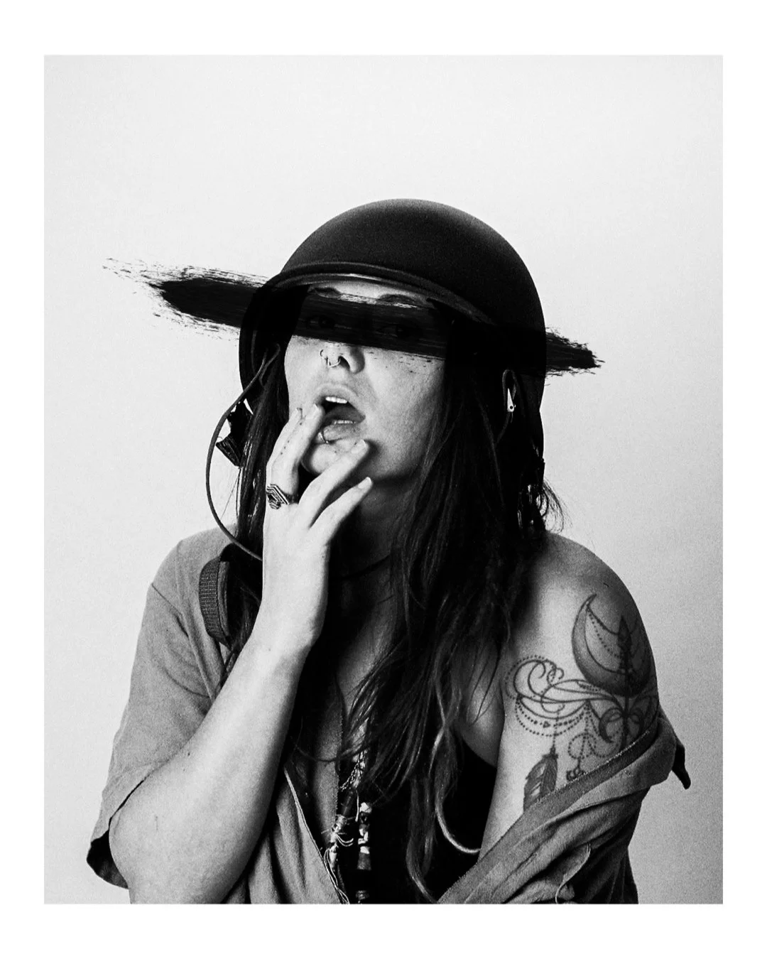 Black and white photo of a woman wearing a helmet with a paintbrush stroke across her eyes, touching her lips, with tattoos on her arm and wearing headphones.