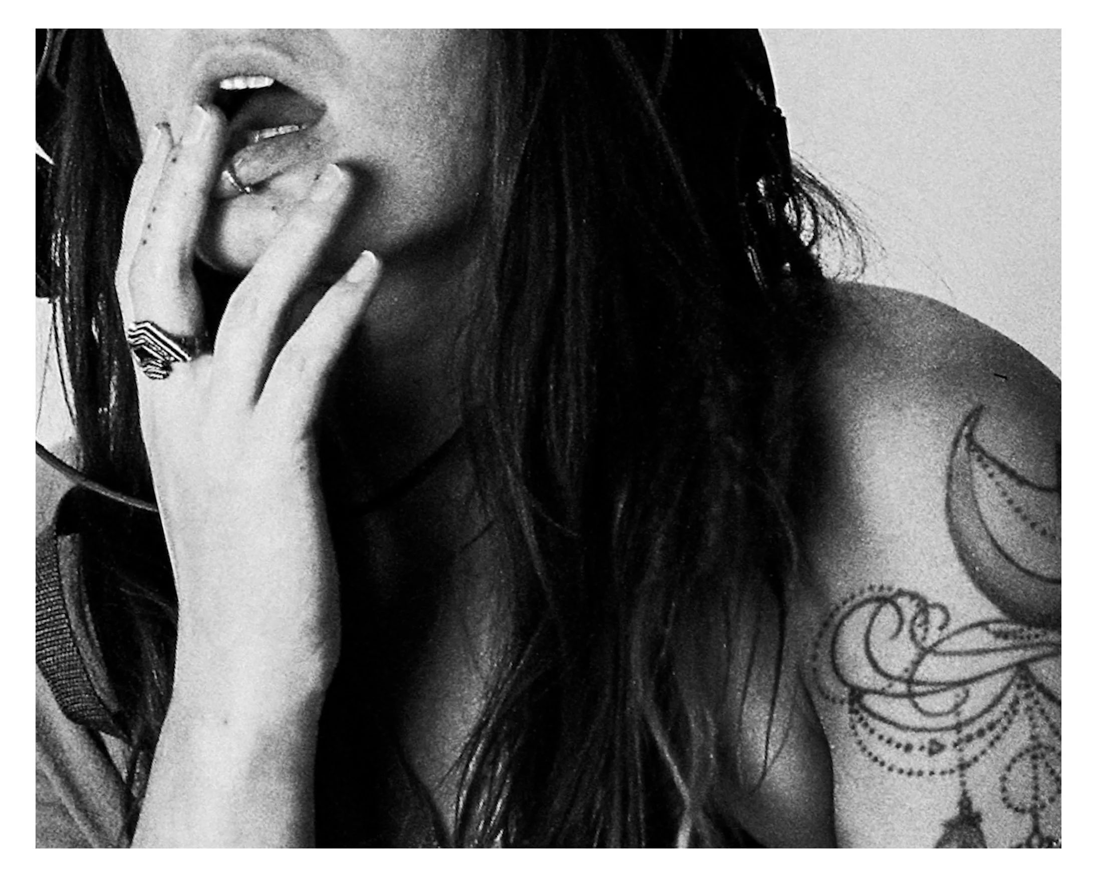 A close-up black and white photo of a woman with long dark hair partially covering her face. She is touching her lips with one hand, which has a ring on the middle finger. She has a visible tattoo on her shoulder.