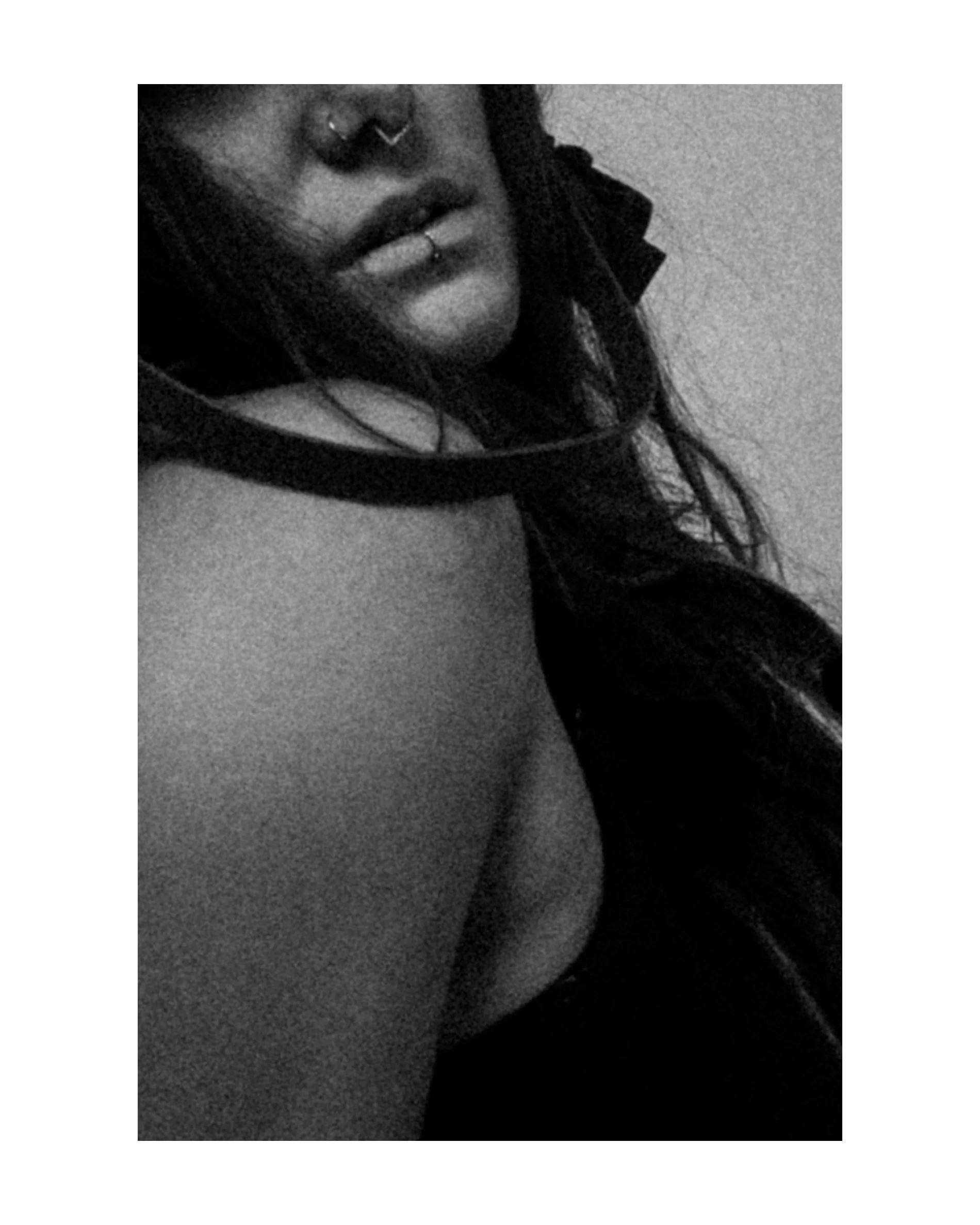 Close-up black and white photo of a woman with long hair, nose and lip piercings, and part of her face, shoulder, and upper chest visible.