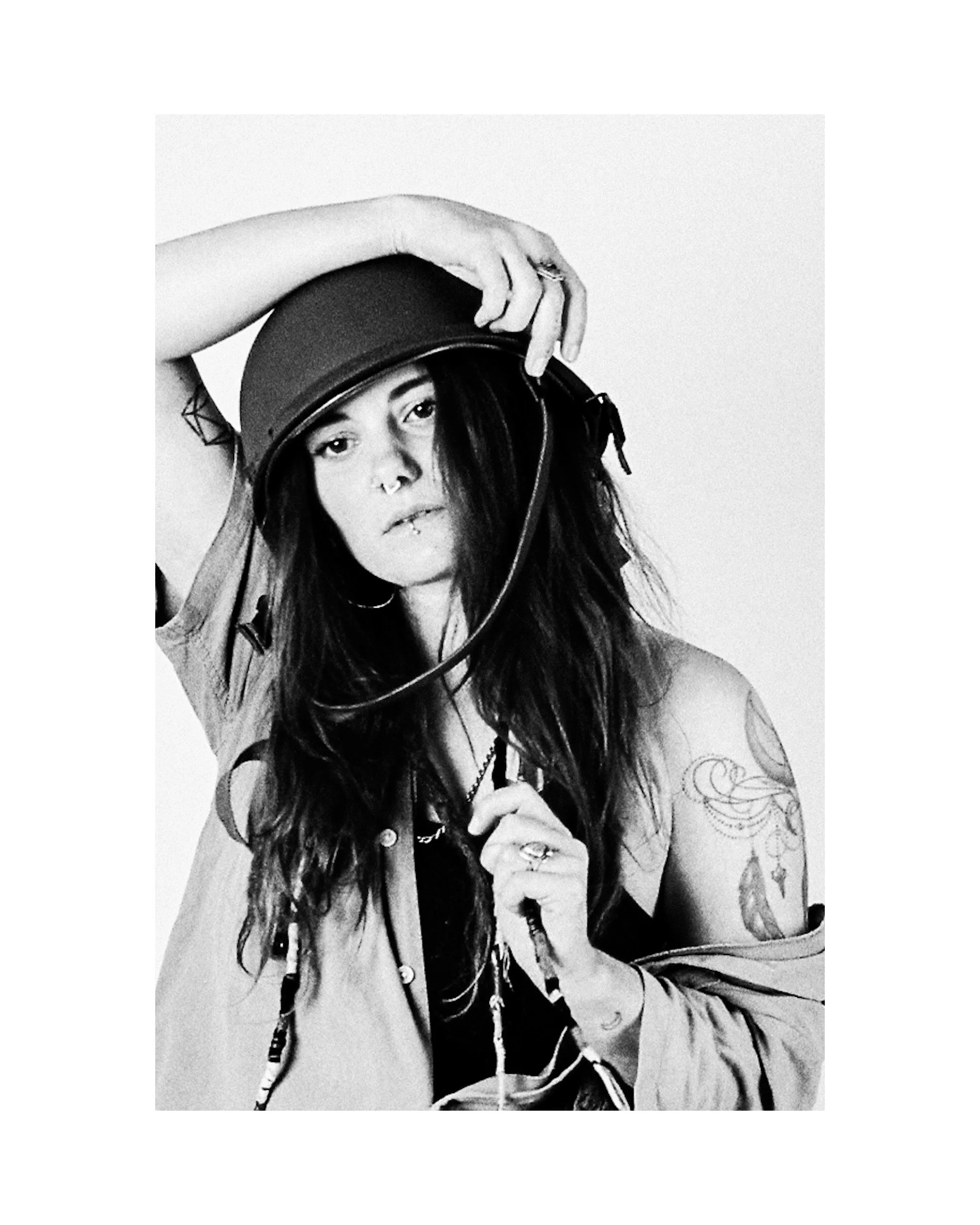 A woman with long hair, tattoos, and piercing holds a helmet and looks at the camera in a black and white photo.