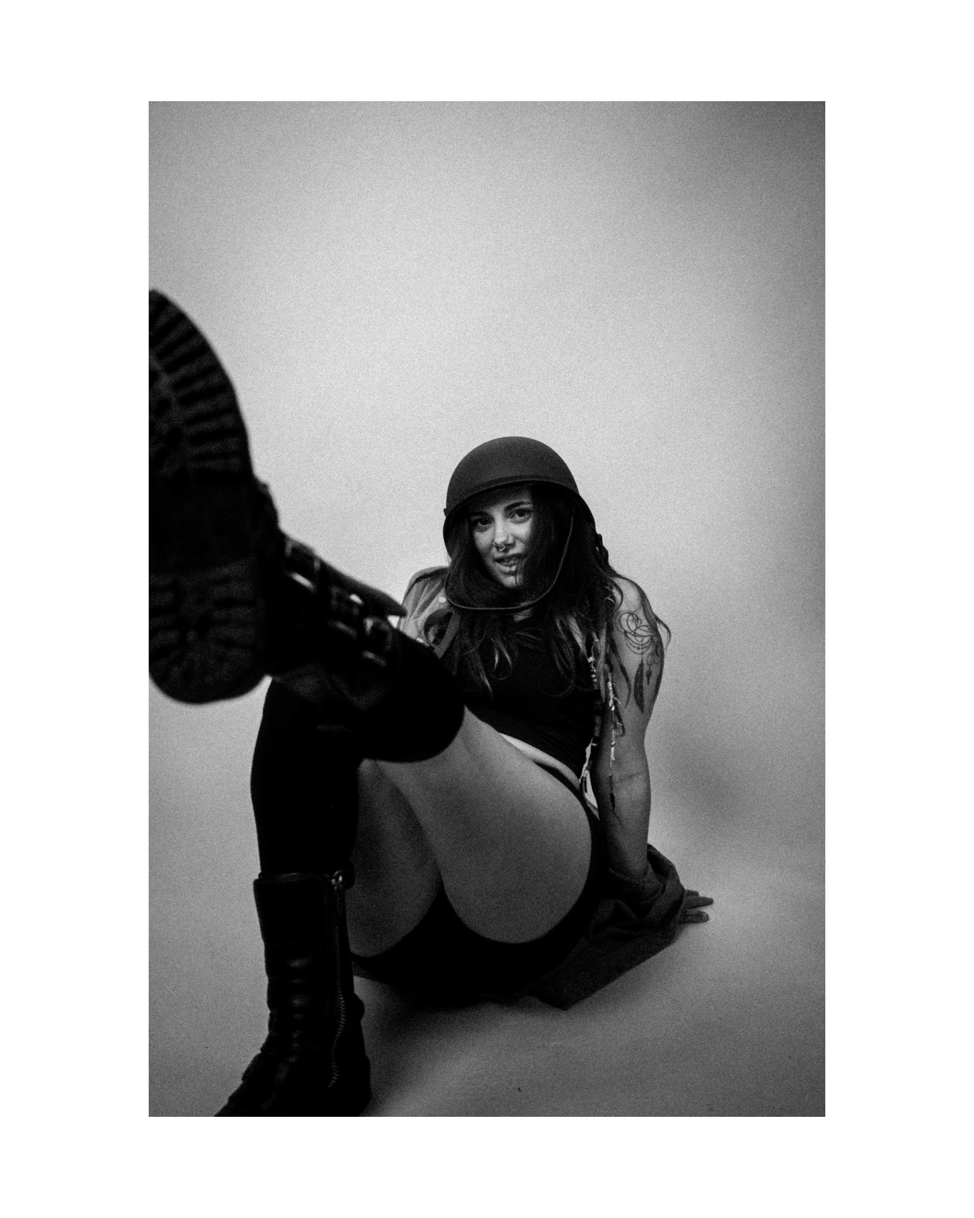 Black and white photo of a woman sitting on the floor, wearing a helmet, leather jacket, boots, and shorts, with tattoos visible on her arm. She is looking at the camera with a confident expression.