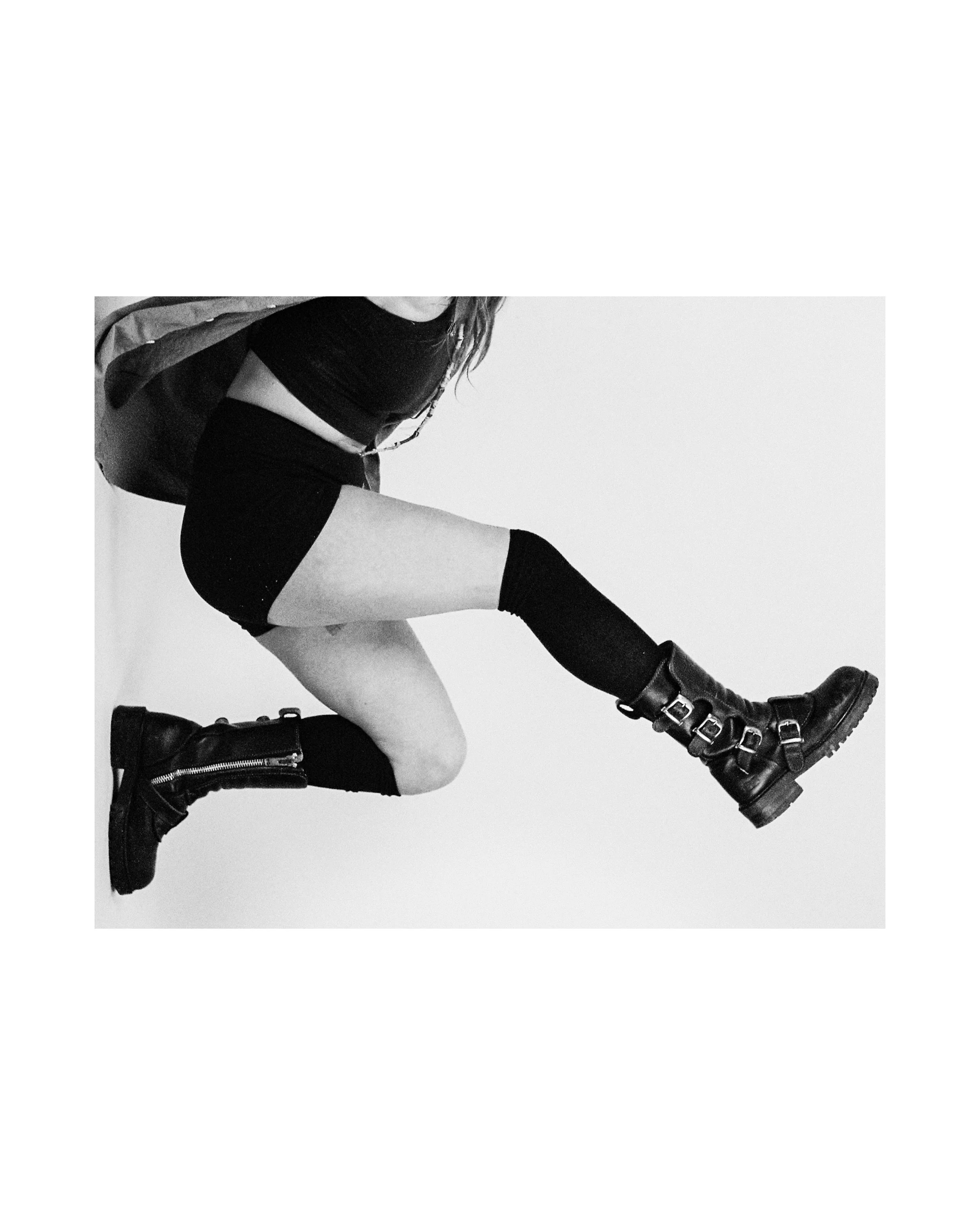A woman wearing a black crop top, black shorts, thigh-high socks, and combat boots, mid-jump against a plain background.
