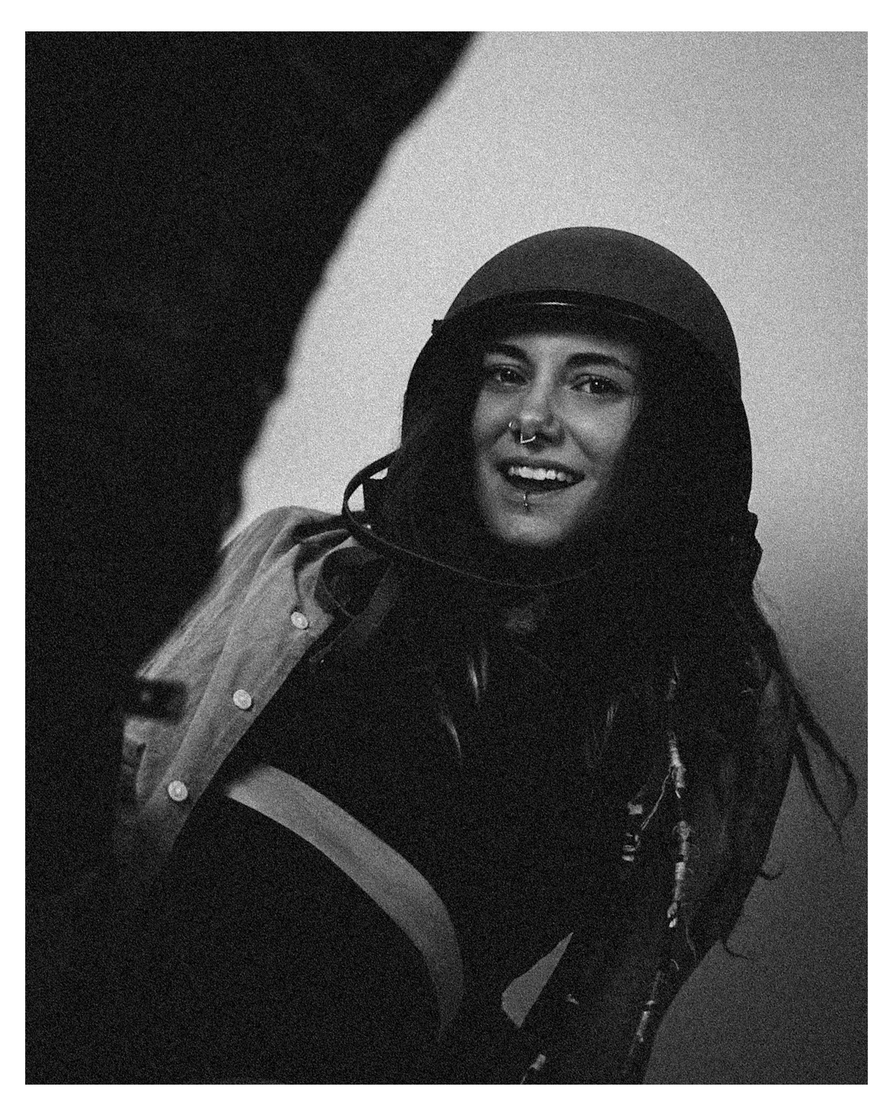 A smiling woman wearing a helmet and a leather jacket, with a nose ring and labret piercing, looking at the camera.