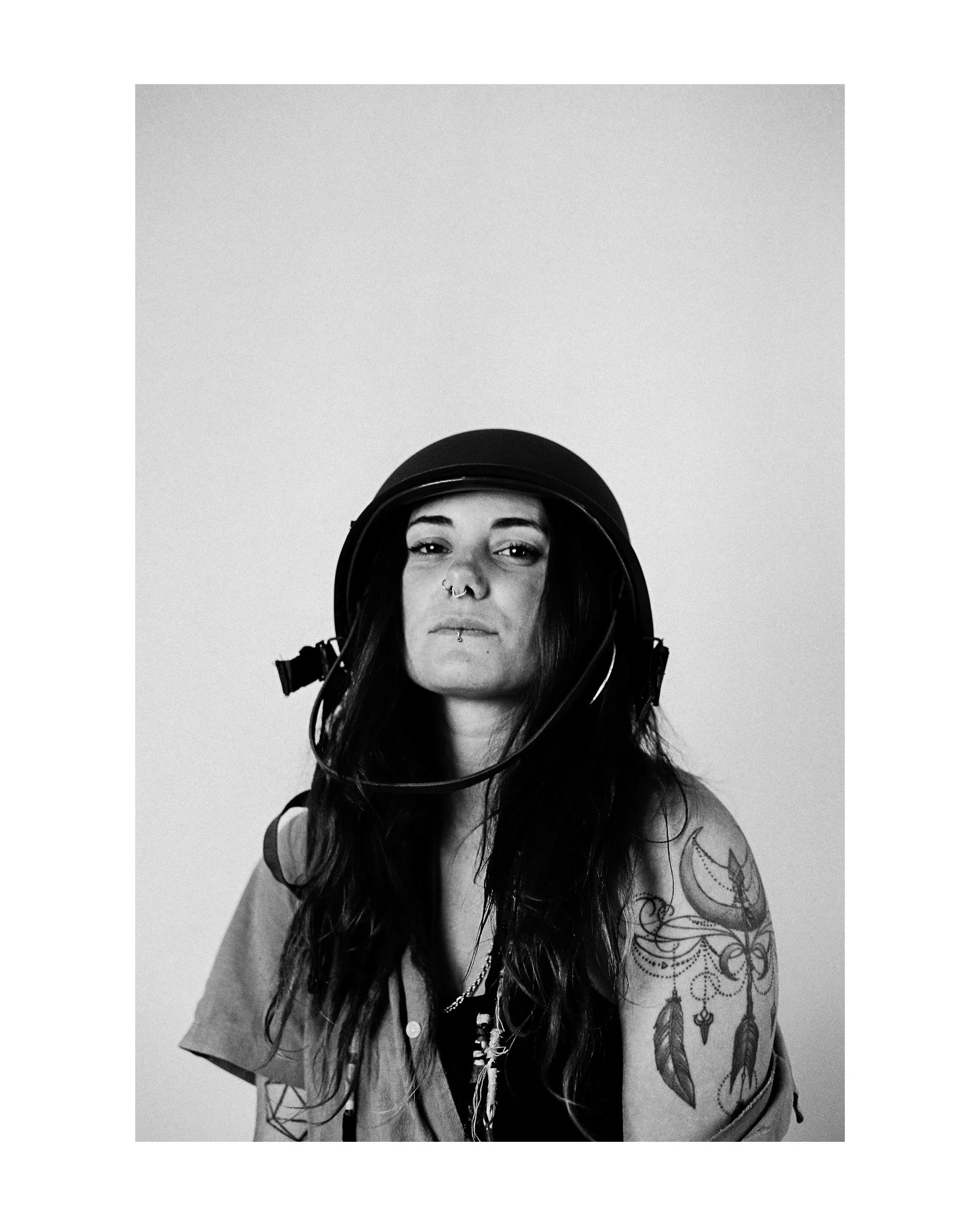 Black and white photo of a woman wearing a motorcycle helmet, with tattoos visible on her arm, looking directly at the camera.