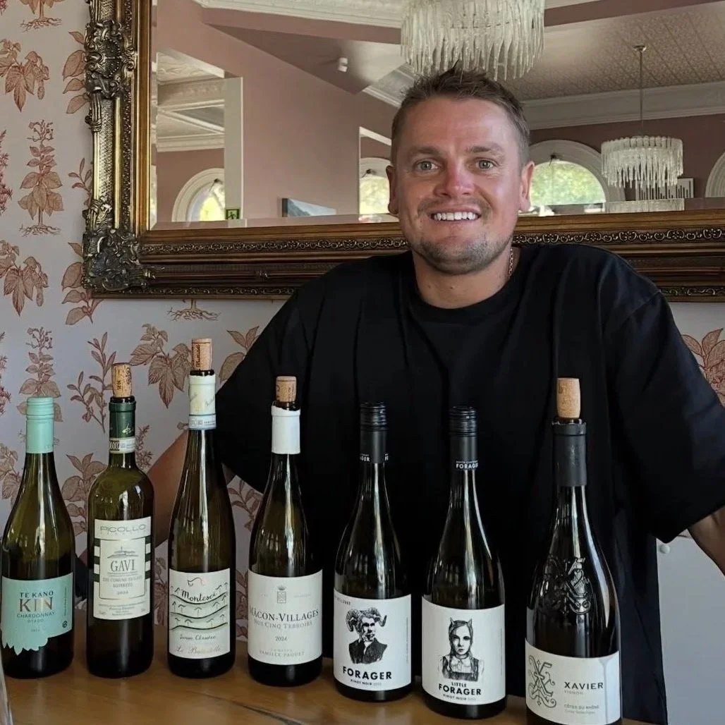 A smiling man sitting behind a table with seven bottles of wine, in a room with a large mirror, floral wallpaper, and elegant chandeliers.