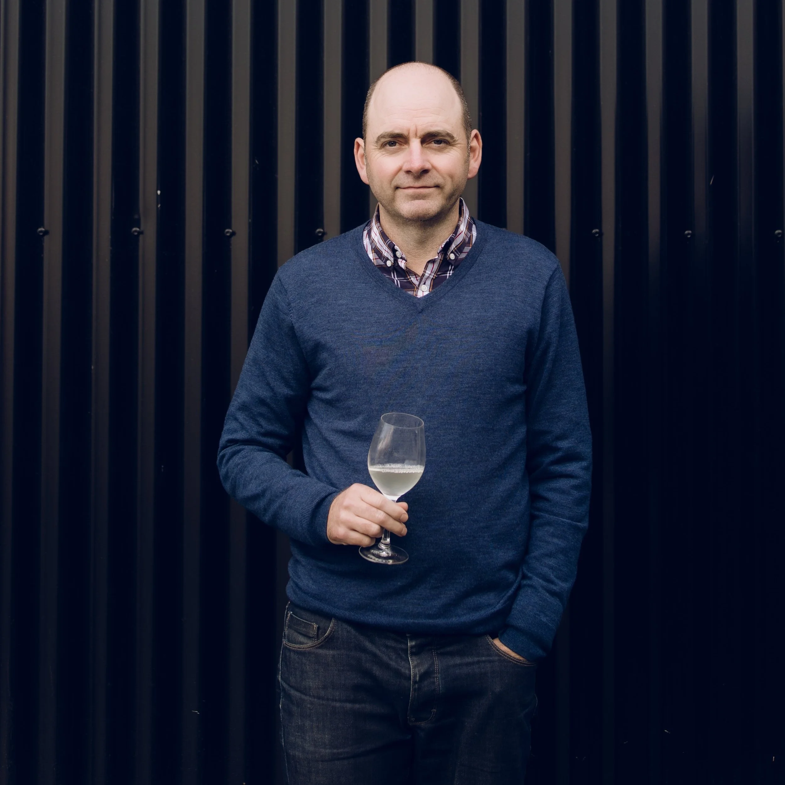 A man with a receding hairline wearing a blue sweater over a checkered shirt, holding a glass of white wine, standing in front of a black vertical panel background.