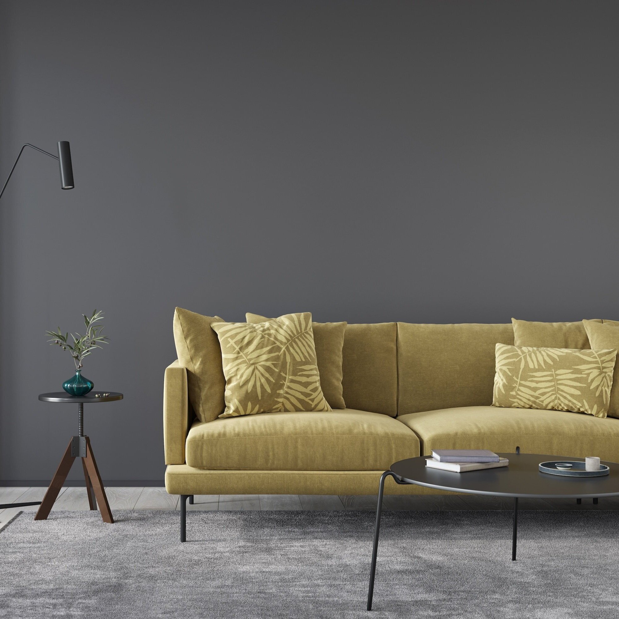 Modern living room with a yellow sofa, accent pillows featuring tropical leaf patterns, a black coffee table with books and decor, a small side table with a blue vase and potted plant, a black wall sconce, and a dark gray wall.