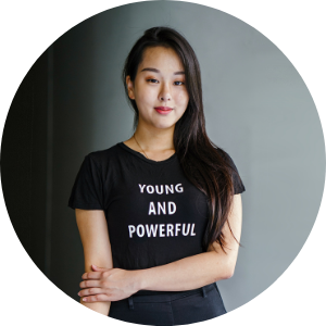 Young woman with long dark hair wearing a black T-shirt that says 'Young and Powerful,' standing against a plain gray background.