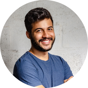 A young man with a beard and dark hair smiling and wearing a blue t-shirt, standing against a concrete wall background.