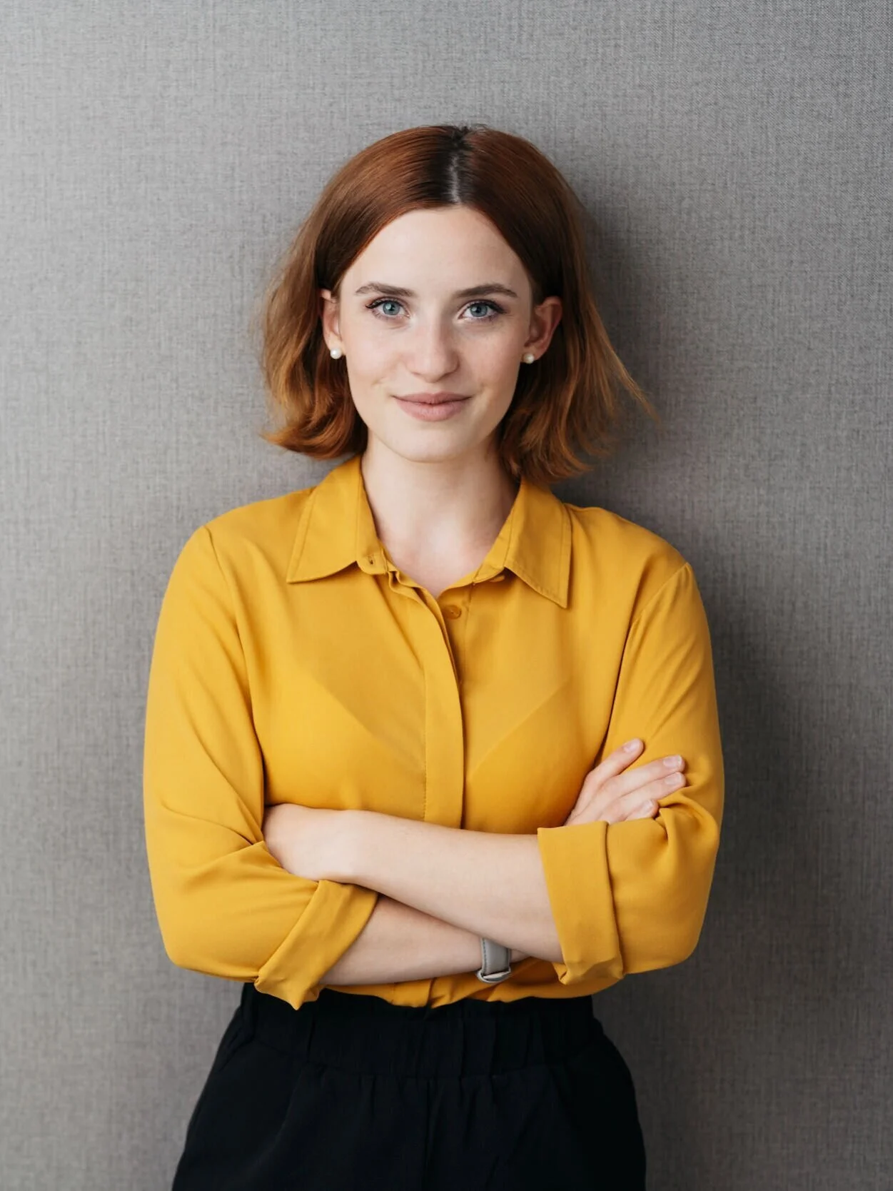 A woman with short reddish-brown hair, wearing a yellow shirt, poses with arms crossed against a gray background.