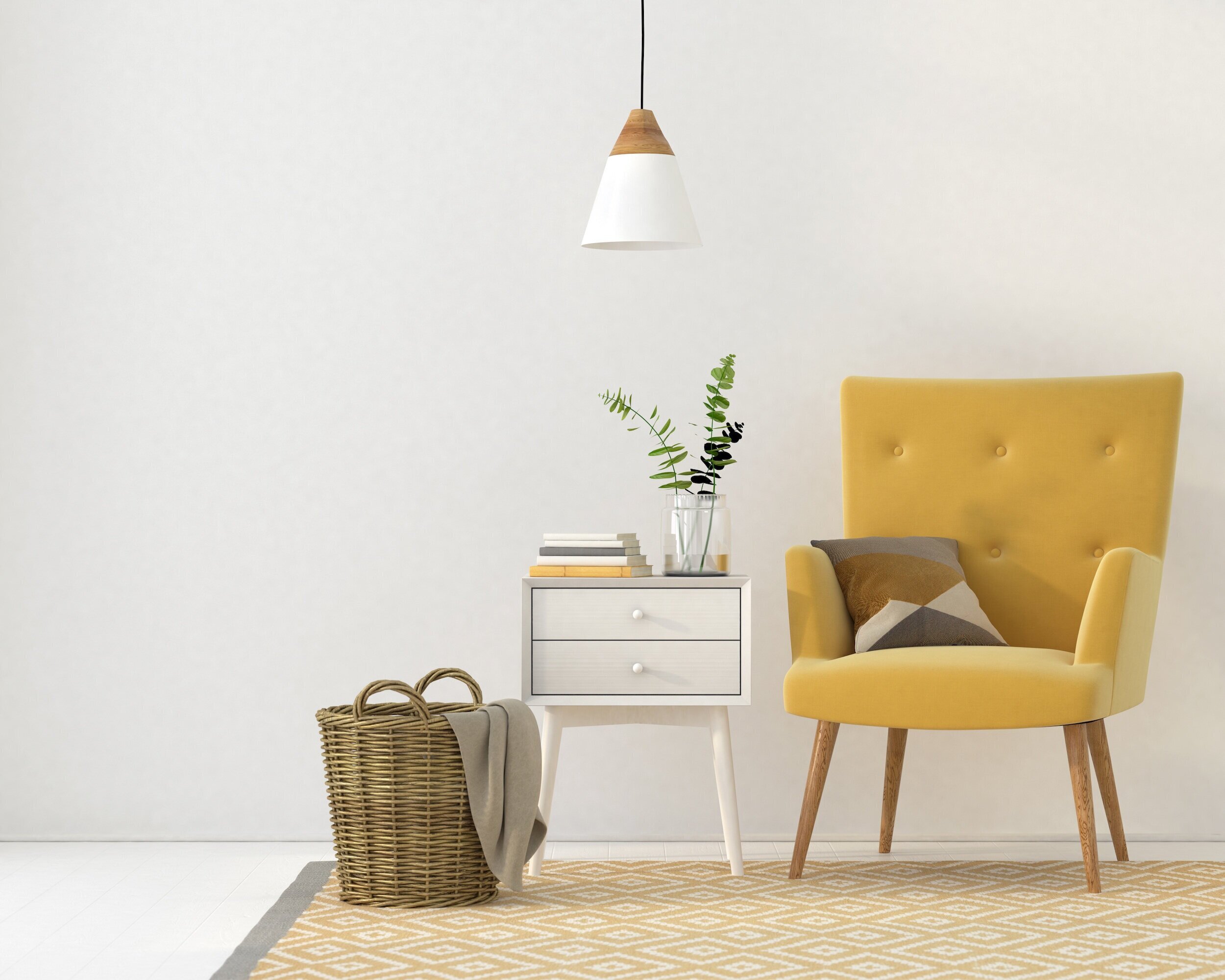 Minimalist interior scene with a yellow armchair, a small white side table with a stack of books, a glass vase with green plant, wicker basket, and a hanging lamp against a plain white wall.