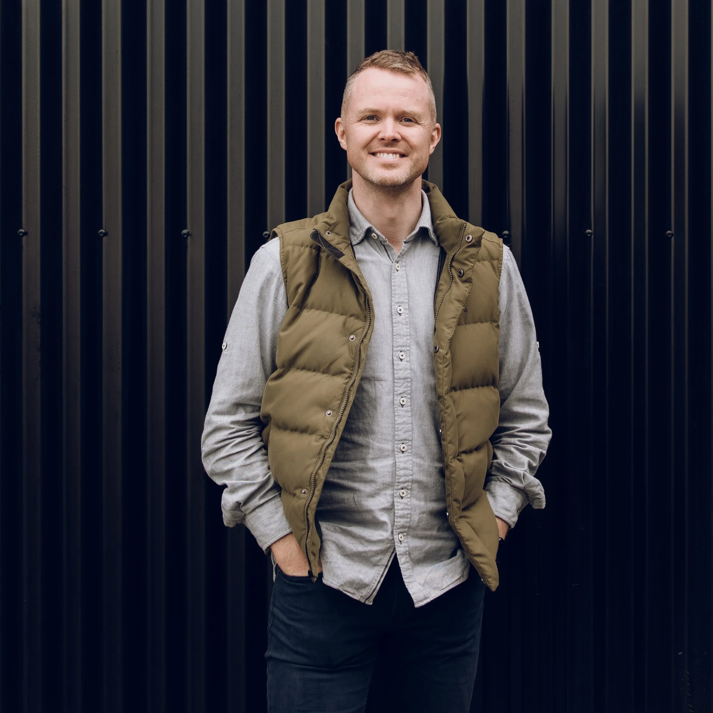 A smiling man with short light brown hair, wearing a light gray shirt and a brown padded vest, standing in front of a black metal fence.