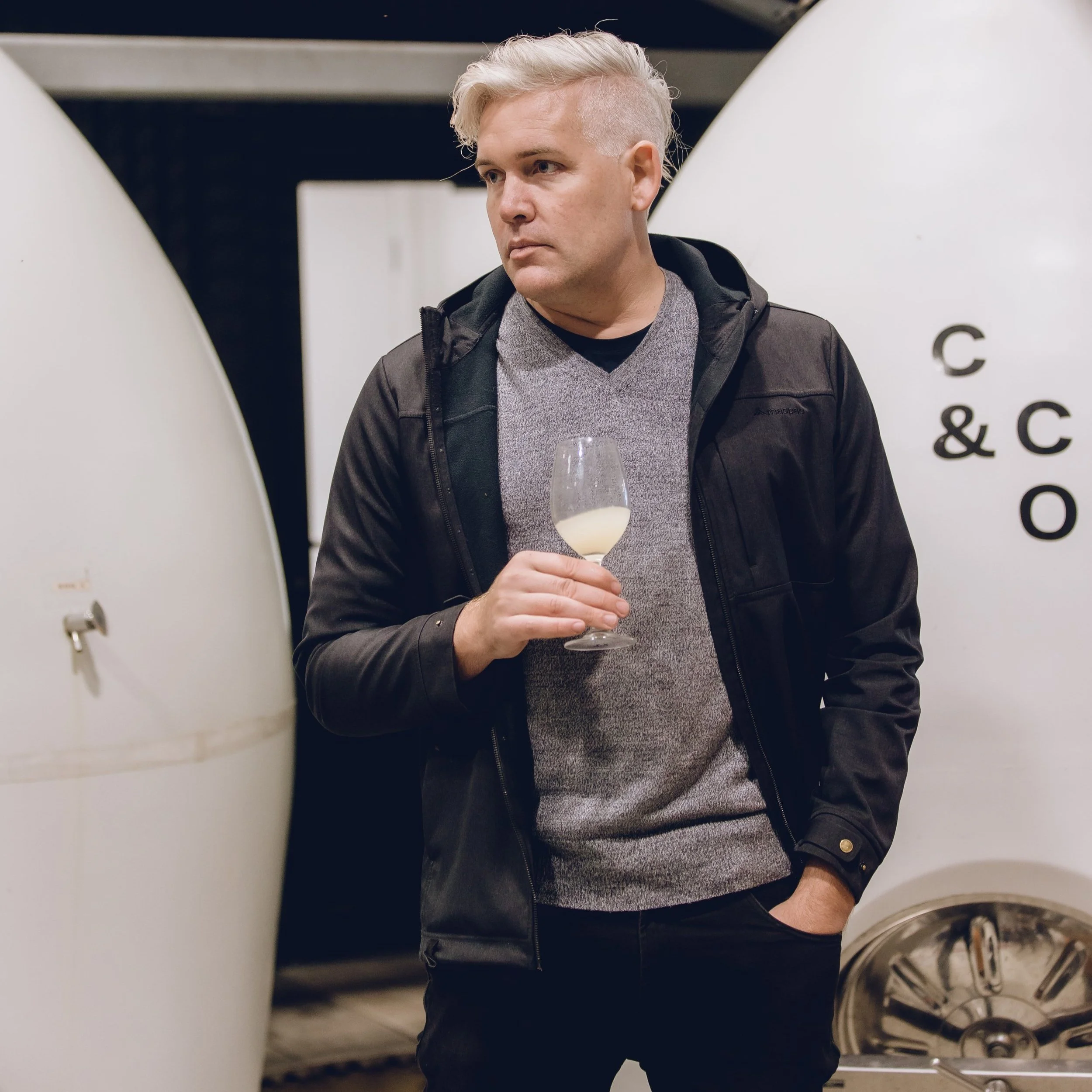 A man with short, platinum blonde hair styled in a pompadour haircut, wearing a dark jacket over a grey sweater, holding a glass of white wine, standing in front of a white wall with black text, next to a commercial washing machine.