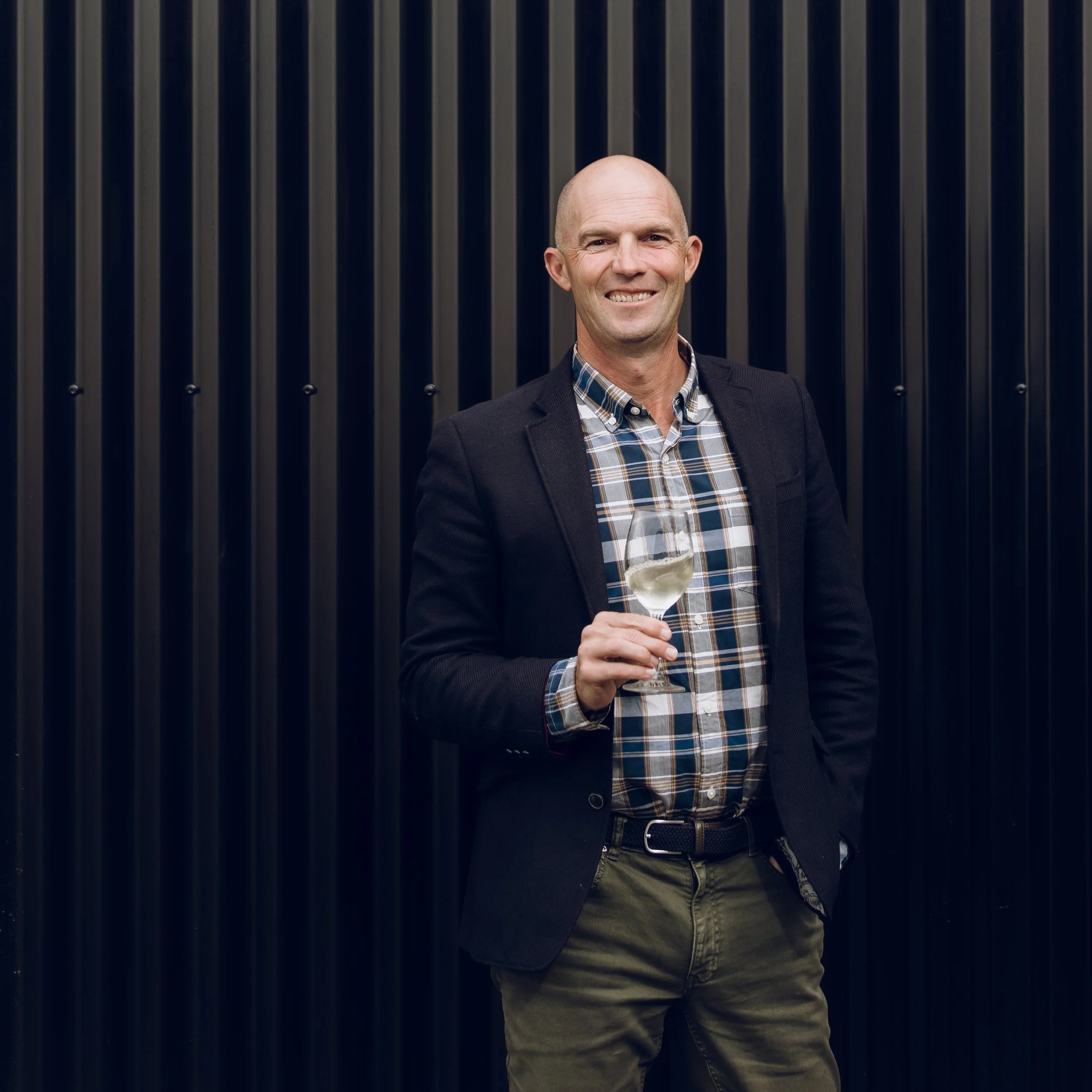A man with a shaved head and a smile, holding a glass of white wine, standing in front of a dark, vertical striped wall, wearing a plaid shirt and a dark blazer.