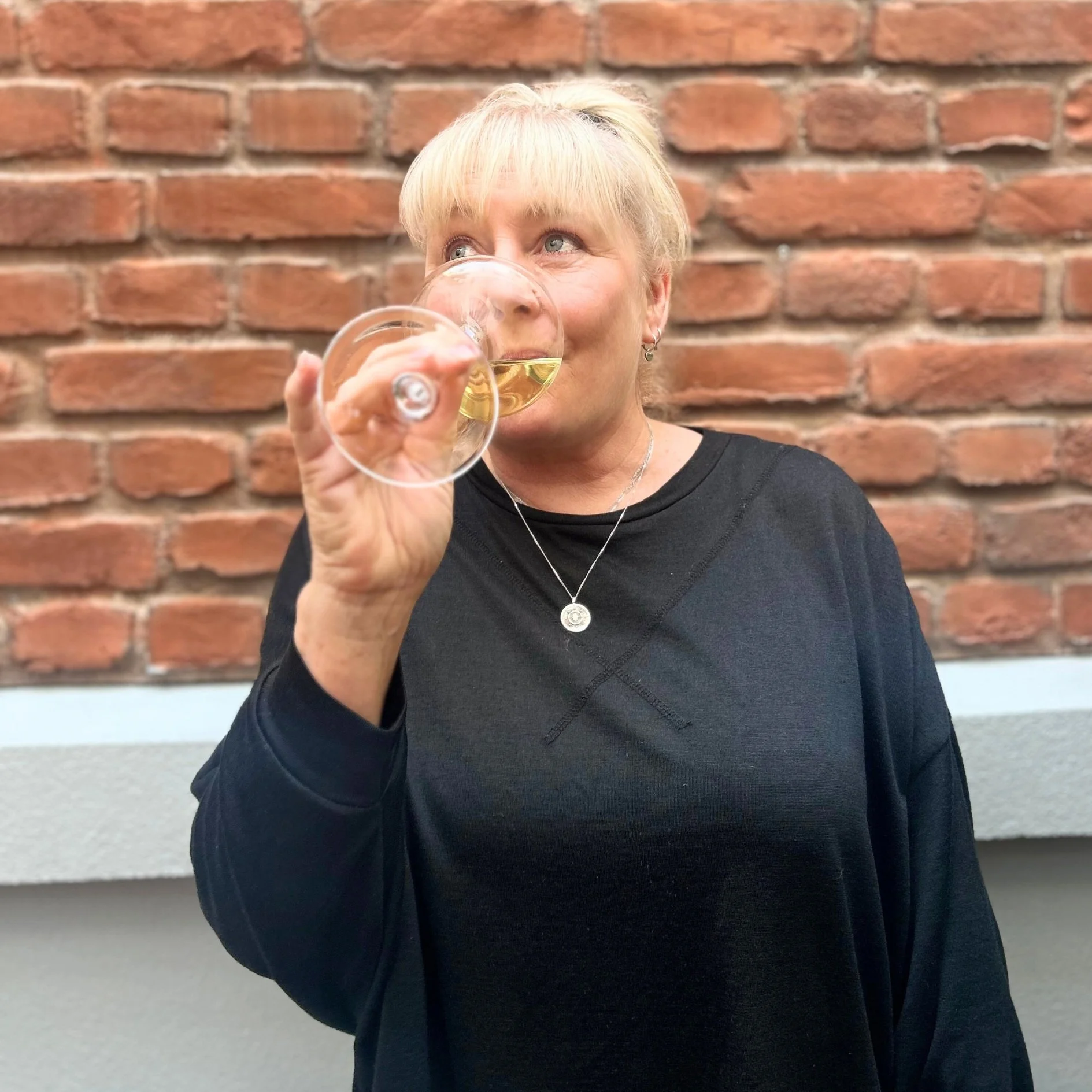 A woman with short blonde hair and earrings taking a sip from a glass of white wine against a red brick wall.