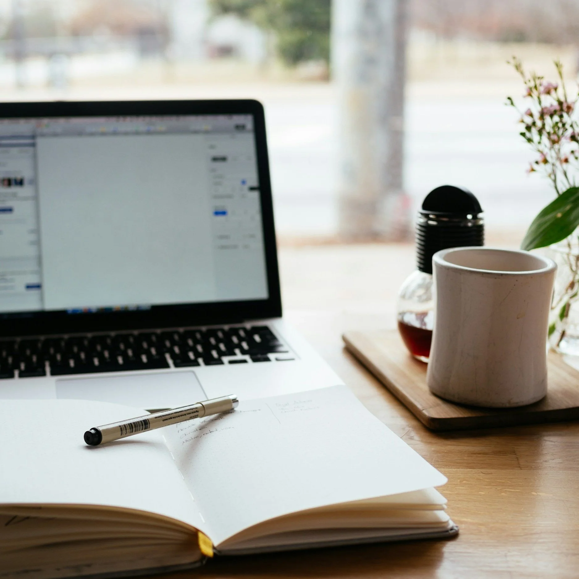 Open notebook with handwritten notes and a pen on a wooden desk, beside a closed book, a white mug, a small plant, and a laptop in a bright room with large windows showing an outdoor view.
