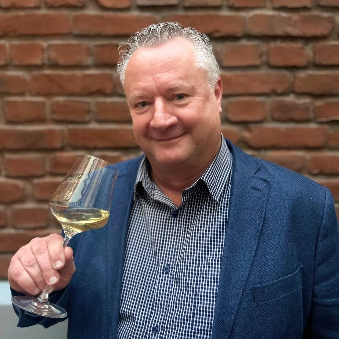 A middle-aged man with short, curly gray hair, smiling, holding a glass of white wine, standing in front of a brick wall, wearing a blue blazer and a checkered shirt.