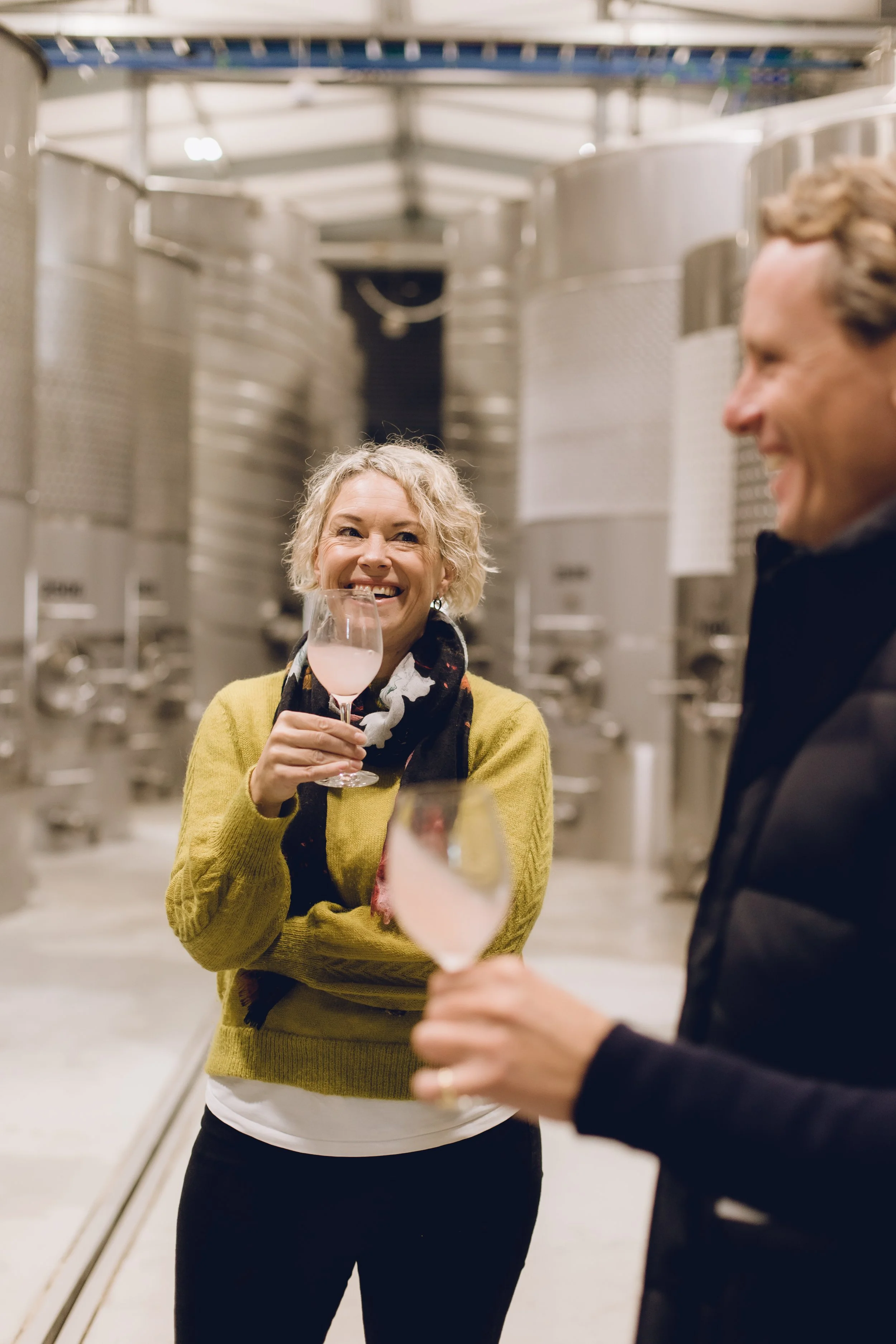 Two people at a winery tasting with large stainless steel fermentation tanks in the background. The woman on the left, with curly blonde hair, is smiling and holding a glass of pink wine. She is wearing a yellow sweater and a patterned scarf. The man on the right, with light brown hair, is smiling and wearing a dark jacket.