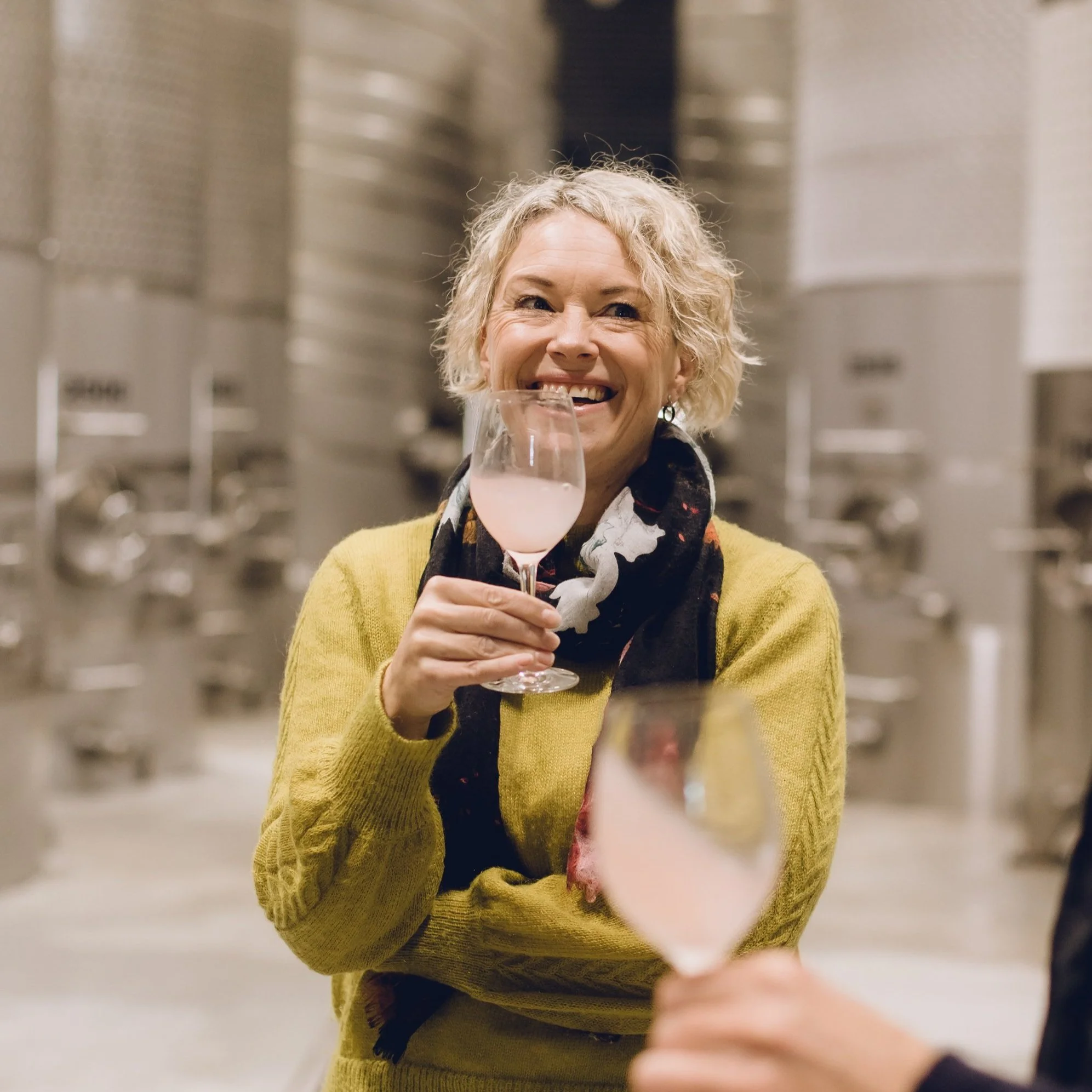 A woman with curly blonde hair smiling and holding a glass of light pink liquid in a wine glass, wearing a yellow sweater.