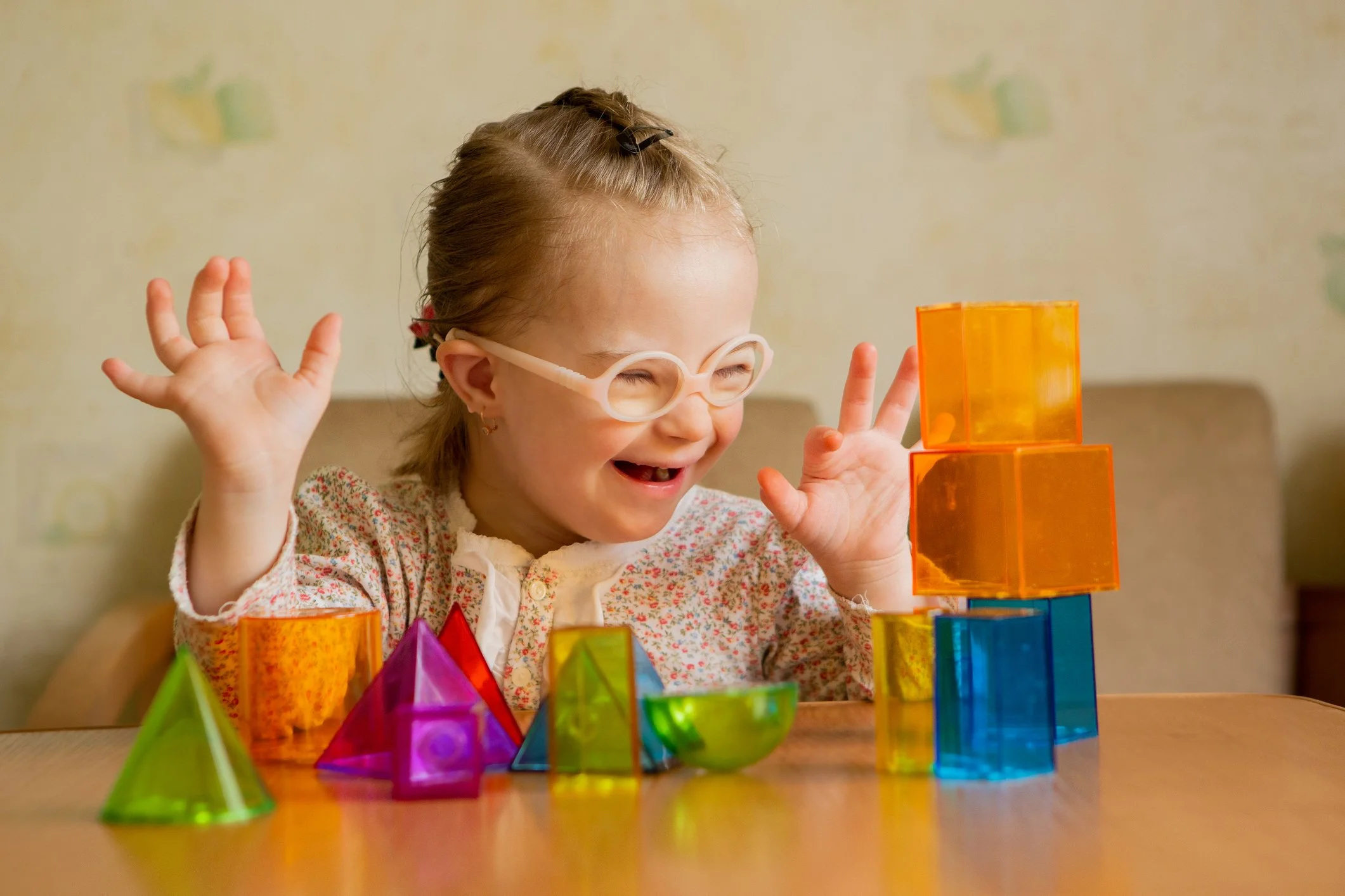 A young girl with glasses and a floral shirt playing with colorful transparent geometric blocks, smiling and raising her hands.