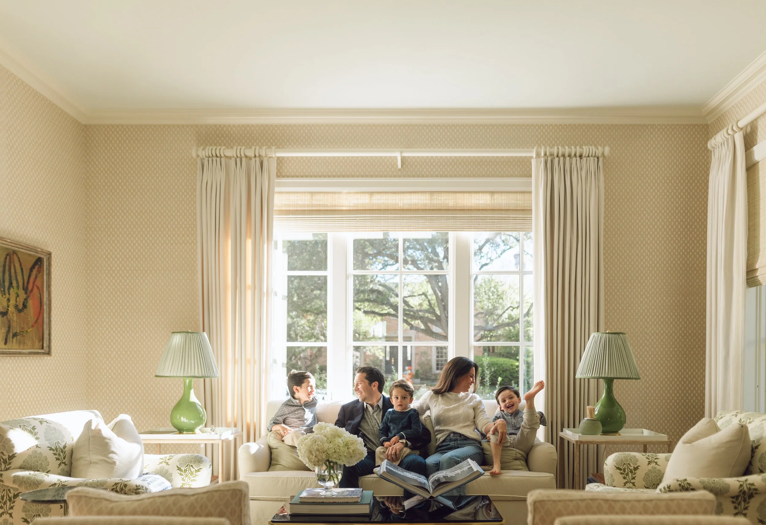 Family of six sitting on a beige couch in a living room with large window and green lamps, enjoying quality time together.