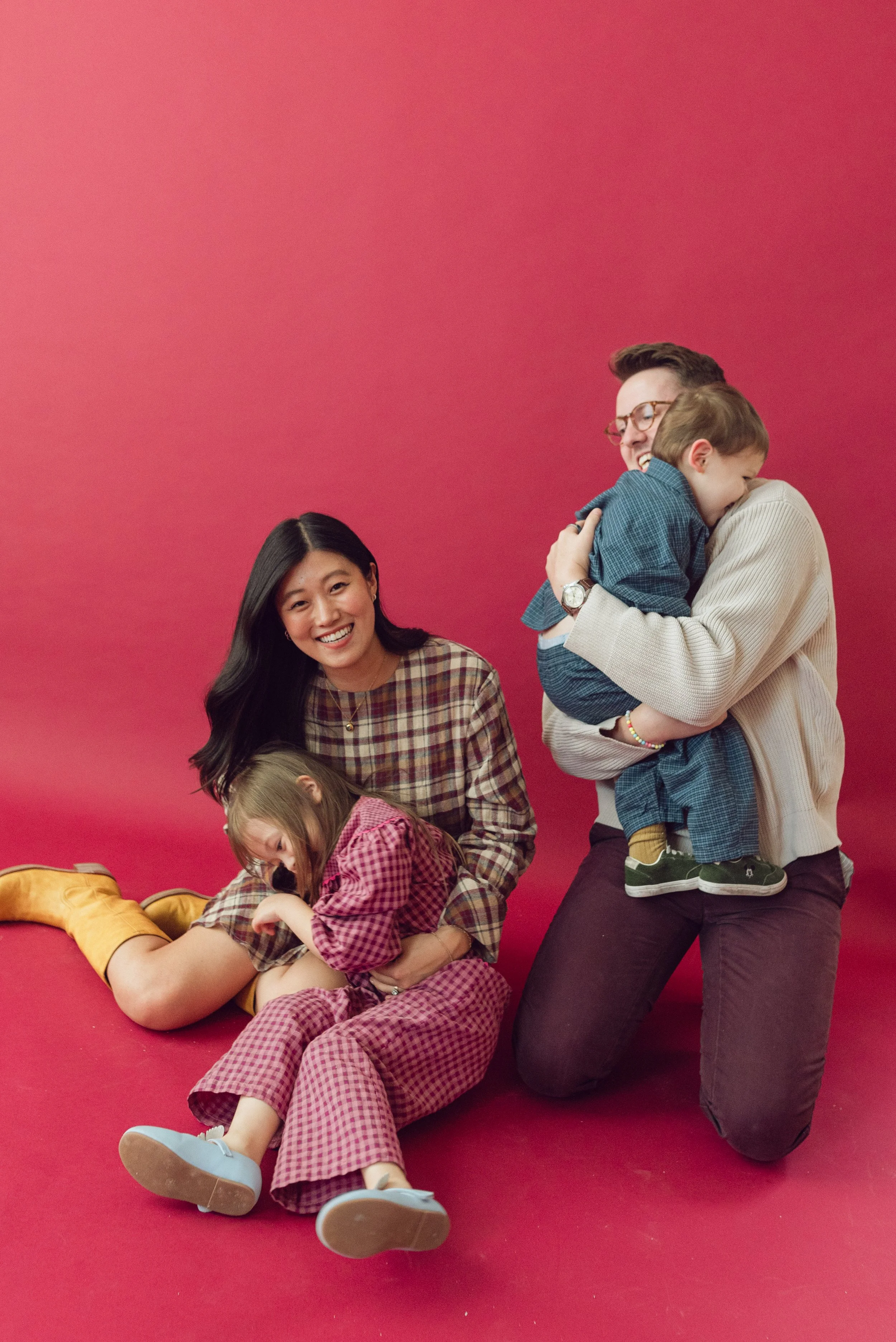A group of four people, including two children, is laughing and hugging each other in front of a red backdrop. The woman is sitting on the floor, the girl is lying on her lap, and the man is kneeling while holding a young boy.