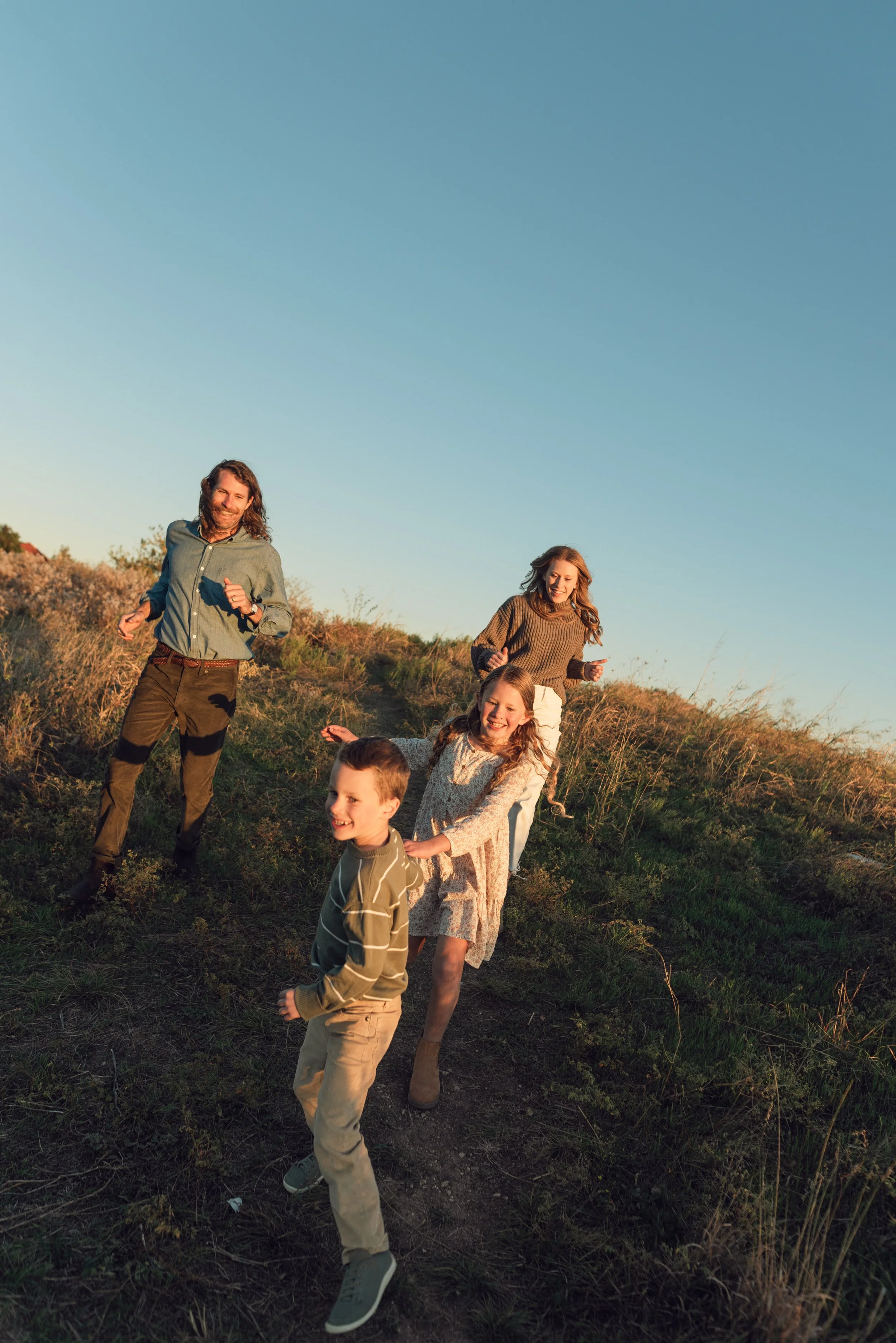 A family of five, including two women and three children, running on a grassy hillside during sunset.