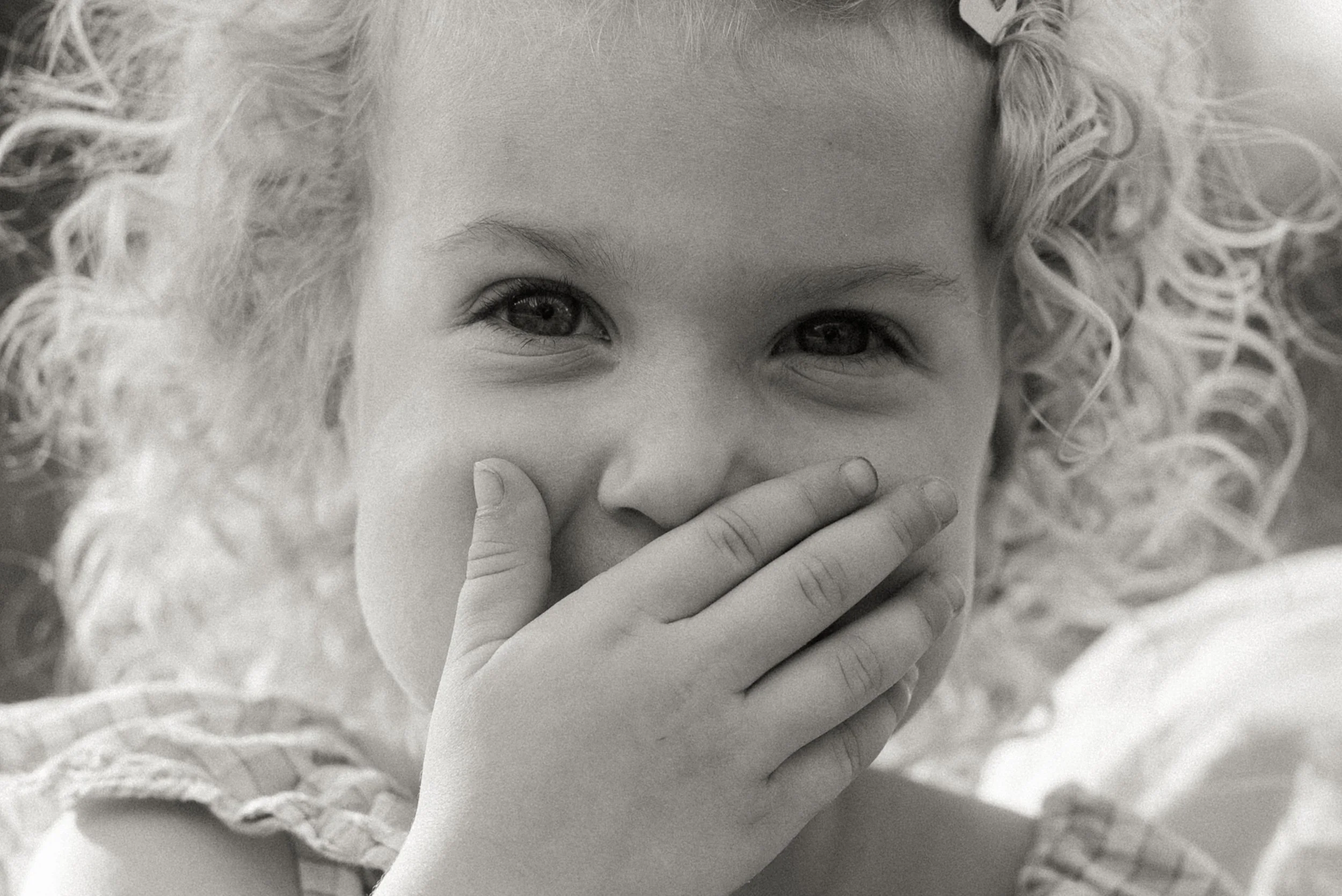 A young girl with curly hair covering her mouth with her hand, smiling with shining eyes.