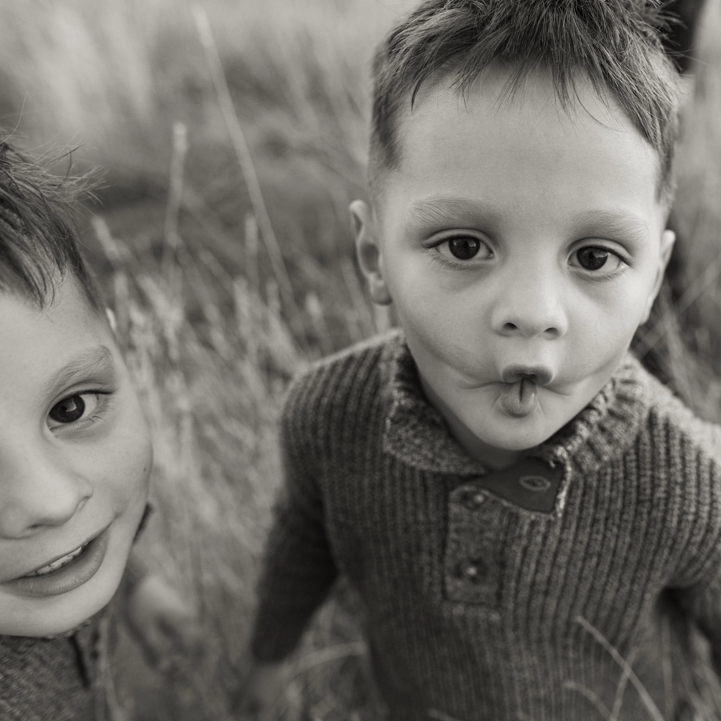 Two young boys outdoors in tall grass, one with finger near mouth, both looking at camera in black and white.