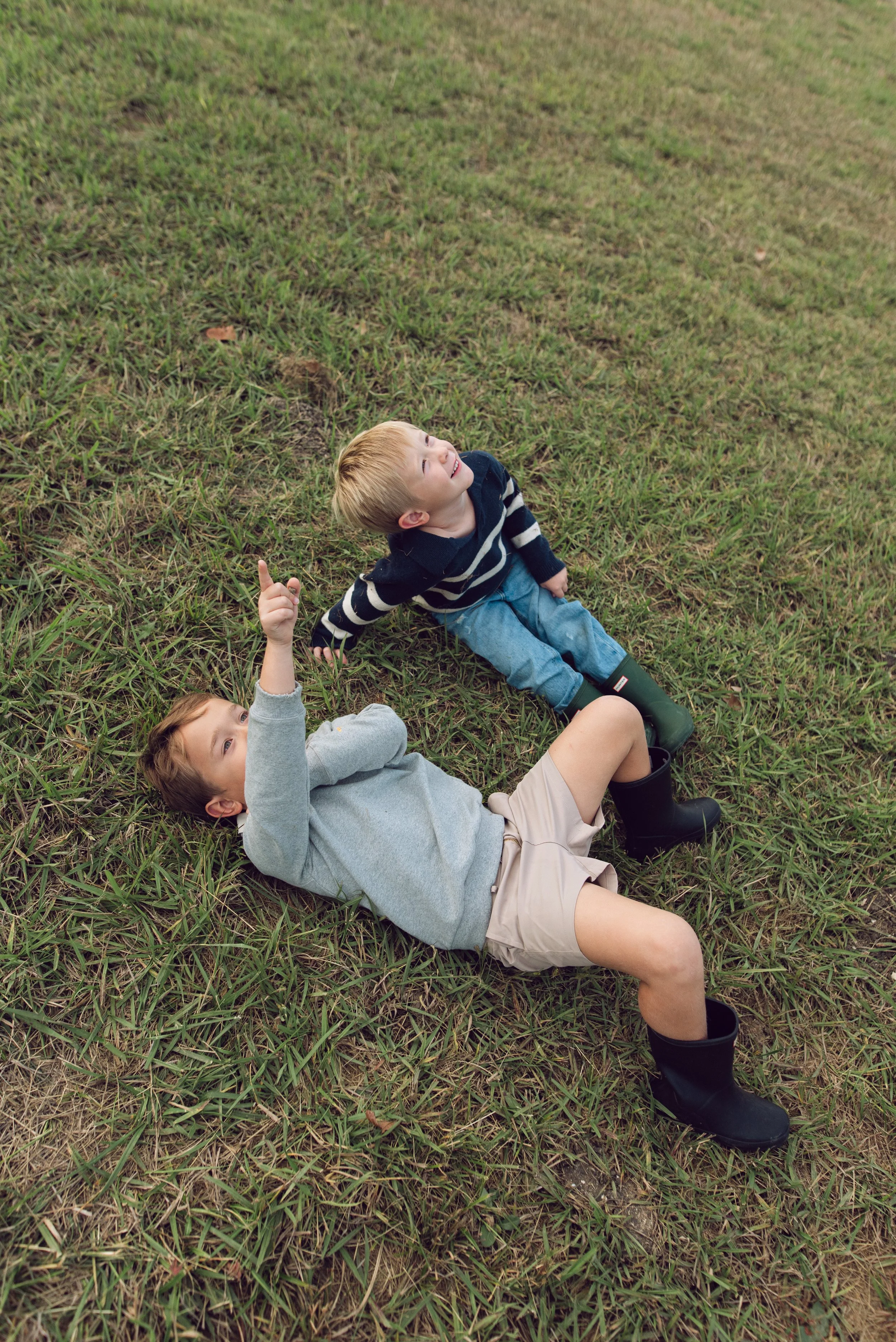 Two young boys lying on grass, looking up and pointing at the sky, with one boy wearing a gray sweatshirt and beige shorts, and the other wearing a navy striped sweater and jeans, both wearing rain boots.
