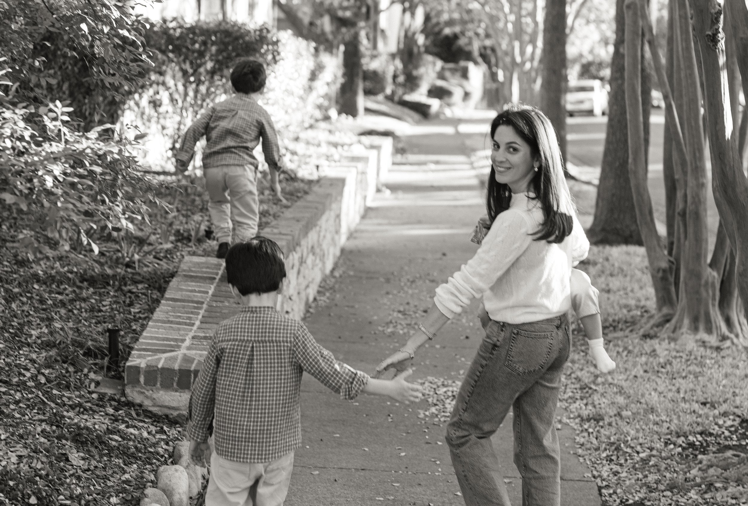 A woman with dark hair smiling and holding hands with a young boy on a sidewalk, while two other children walk ahead on a raised brick platform in a park with trees and bushes.