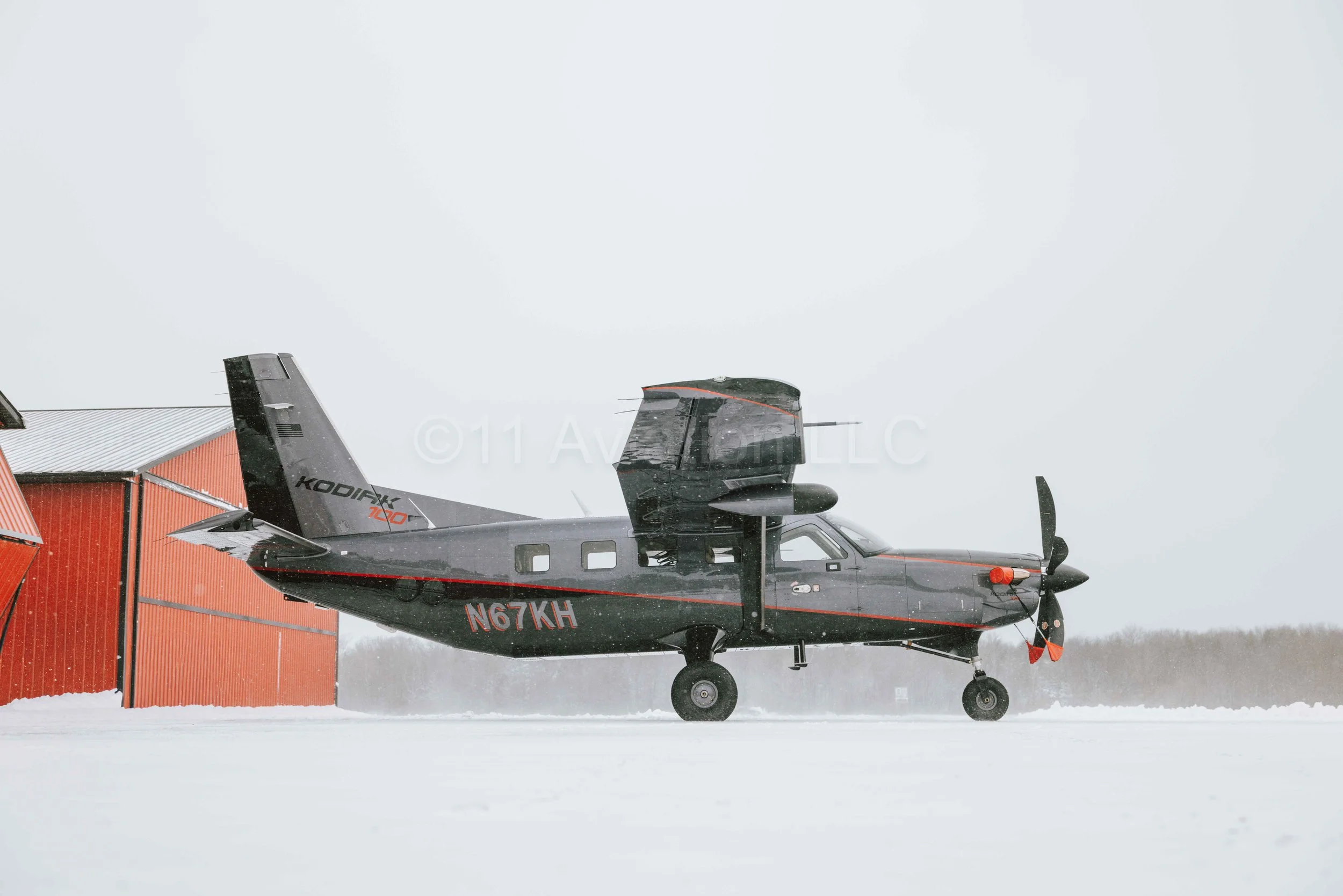 Black Daher Kodiak 100 with red accents parked on snowy ground near red hangar during overcast weather.
