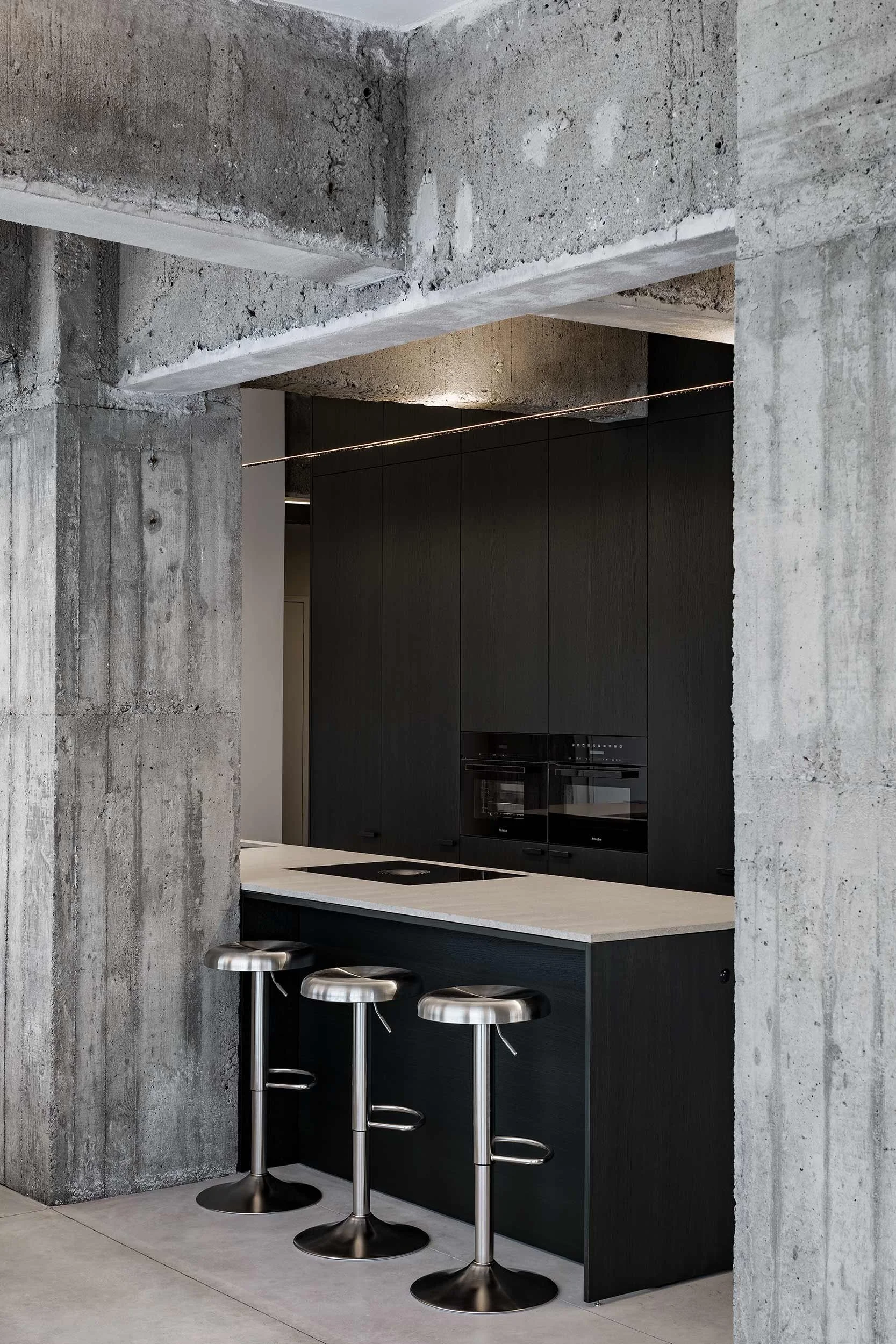 Modern kitchen with black cabinets and a white countertop, concrete walls, and three silver bar stools at a black kitchen island.