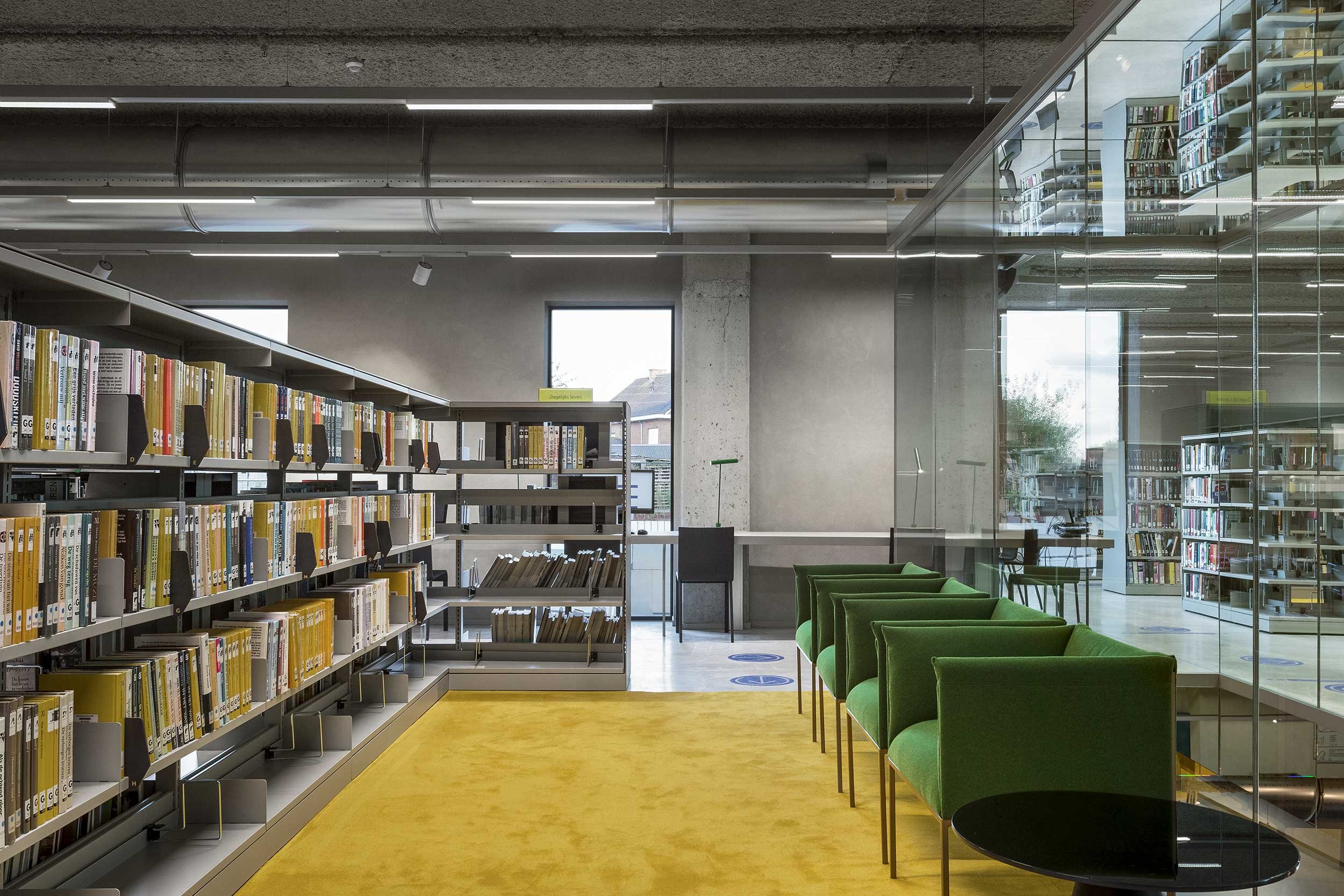 Interno moderno di una biblioteca pubblica con scaffali pieni di libri, sedute verdi, moquette gialla e pareti vetrate che riflettono l’ambiente. Illuminazione industriale a vista e spazi di lettura progettati per il comfort e lo studio.