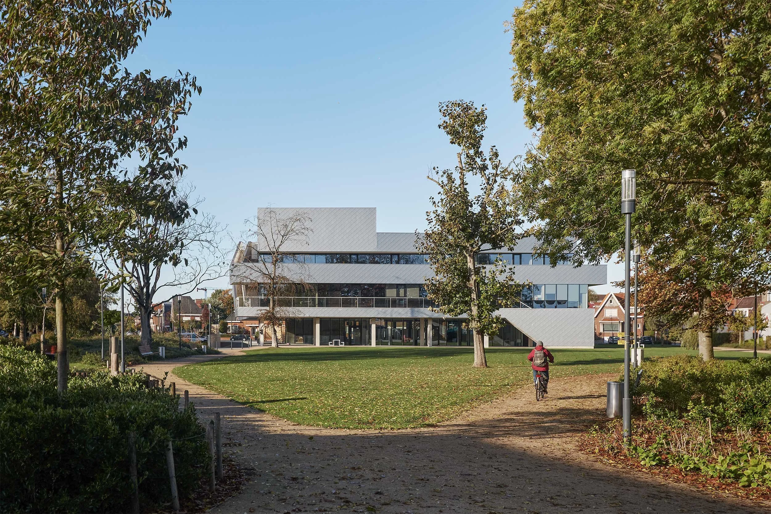 Stadskantoor in Torhout seen from the public park, showing the contemporary civic building integrated into a green landscape with pedestrian paths and trees.