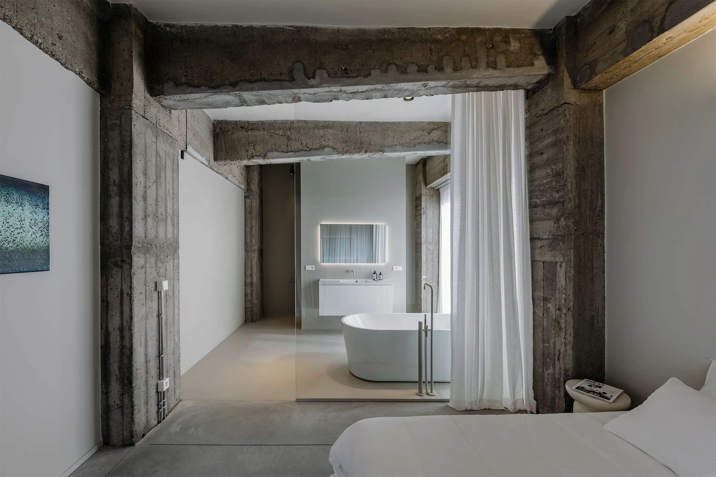 Frontaal zicht op een moderne badkamer met zichtbare betonnen balken en kolommen, een vrijstaand bad en een gedeeltelijk zicht op de aangrenzende slaapkamer.