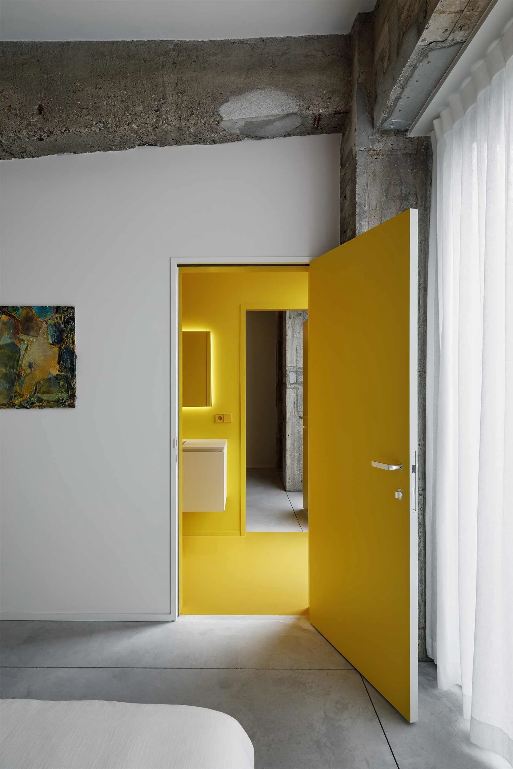 Frontaal zicht vanuit de slaapkamer op een open gele deur die uitzicht biedt op een badkamer met gele wanden, ruwe betonnen kolommen en een industrieel interieur.