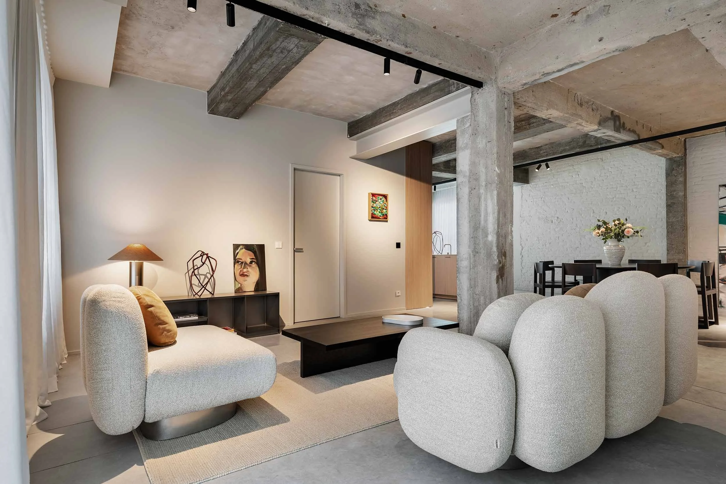 Modern living room with concrete ceilings and exposed beams, cream-colored furniture including rounded armchairs and a sofa, wooden and black coffee tables, a white rug, and artwork including a portrait of a woman and a colorful framed painting. There is a side table with a lamp and a vase with flowers, and a dining area visible in the background.