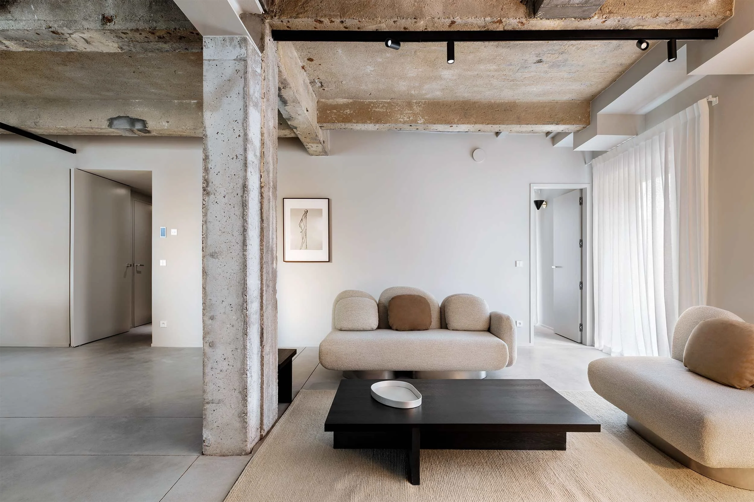 Modern living room with exposed concrete beam and pillar, neutral-colored sofa, black coffee table, framed artwork, white curtains, and track lighting on ceiling.
