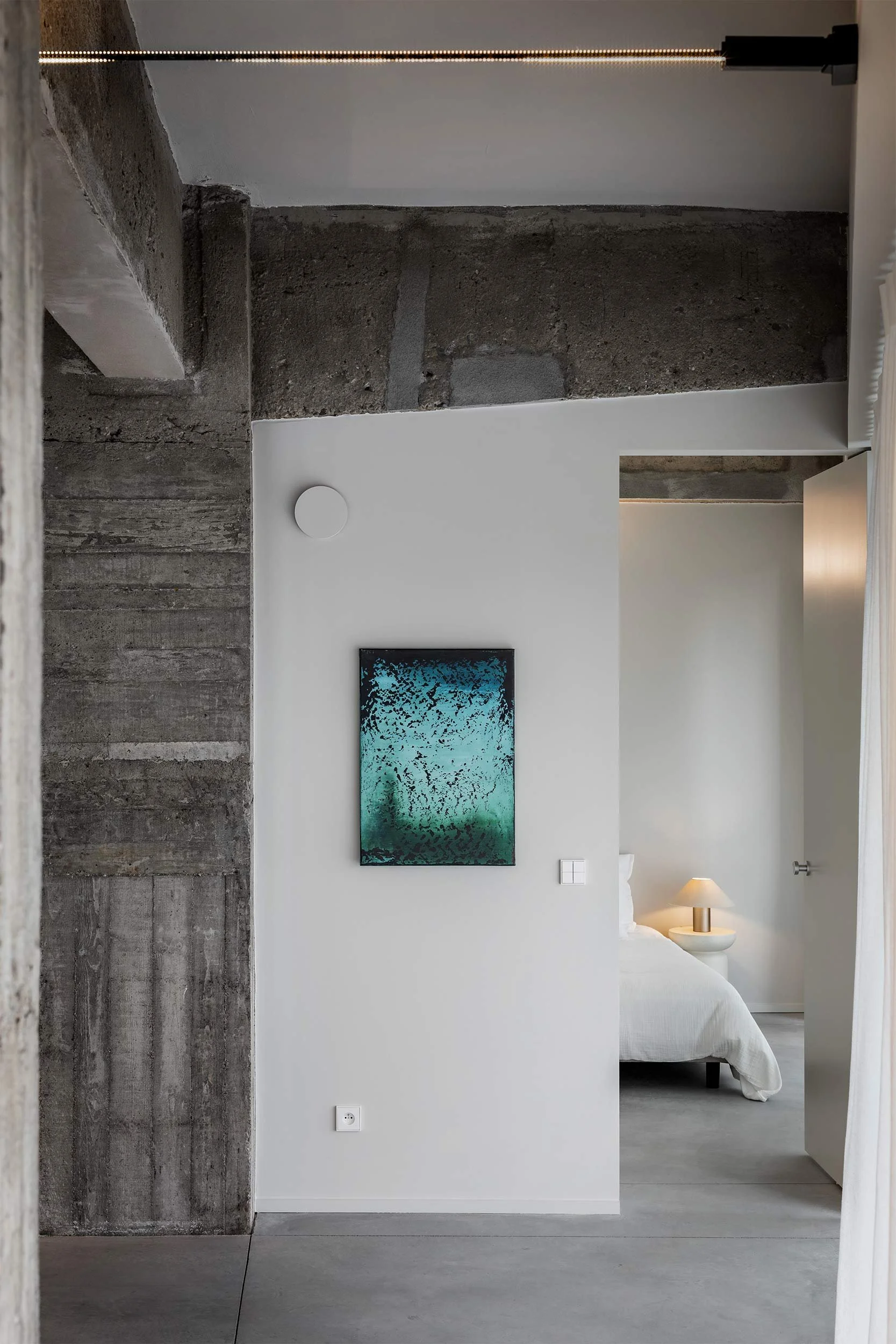 A modern bedroom with a white bed, a bedside table with a lamp, a wall-mounted TV, a painting, and a mix of concrete and white walls.