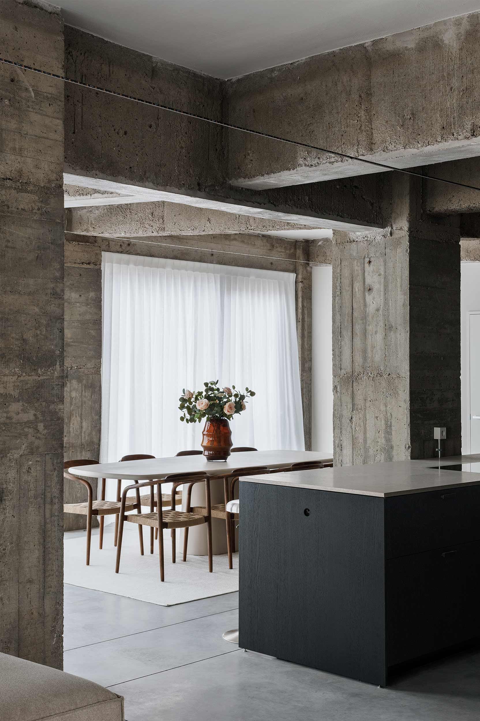 Interior living space with concrete beams and walls, a white dining table with six wooden chairs, a brown vase with pink roses on the table, white curtains over large window, and part of a kitchen counter.
