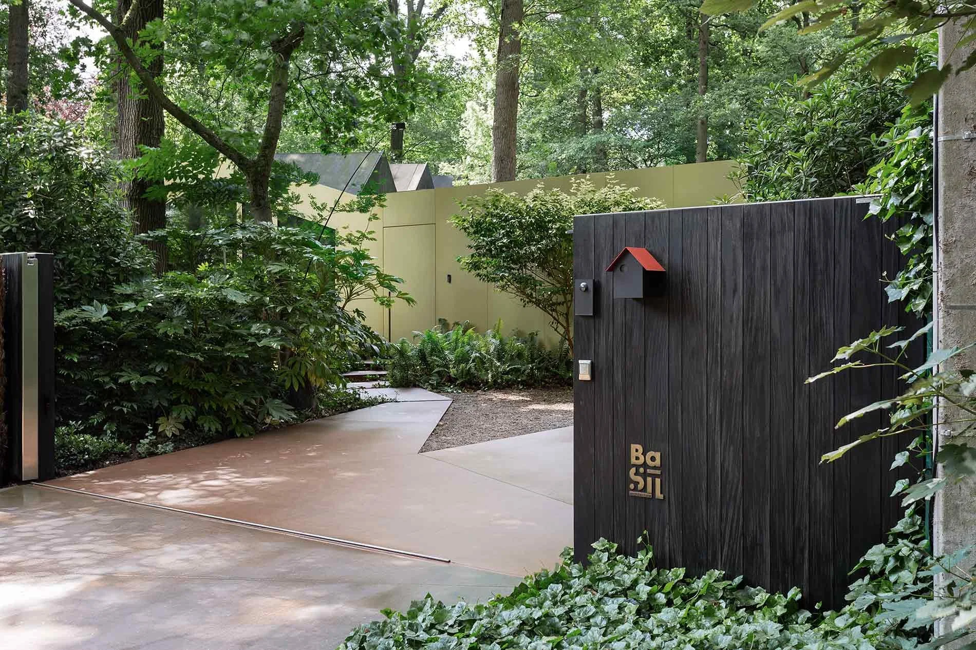 Front yard with a black wooden gate, greenery, trees, shrubs, and a concrete pathway that brings to the entrance of the house in the woods.