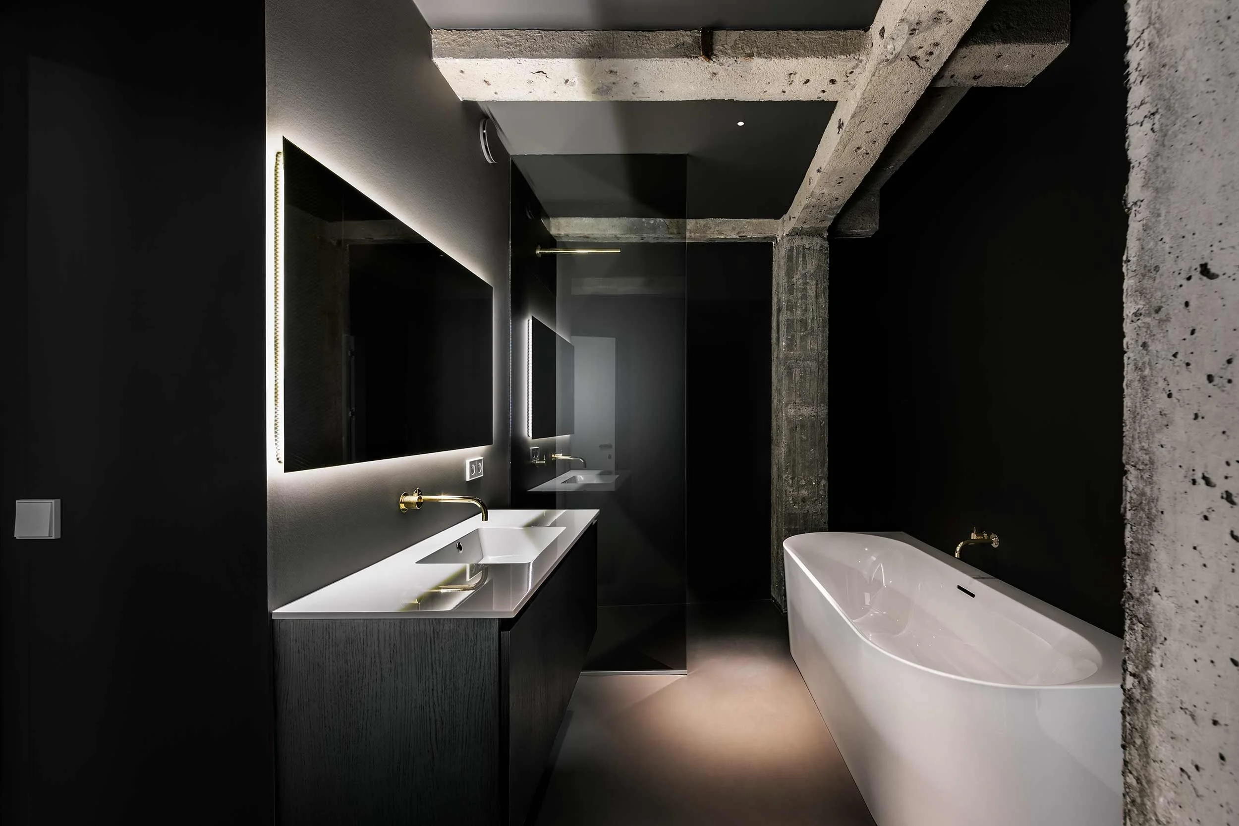 Frontaal zicht op een moderne badkamer met zwarte wanden, betonnen kolommen en gouden VOLA-kranen.