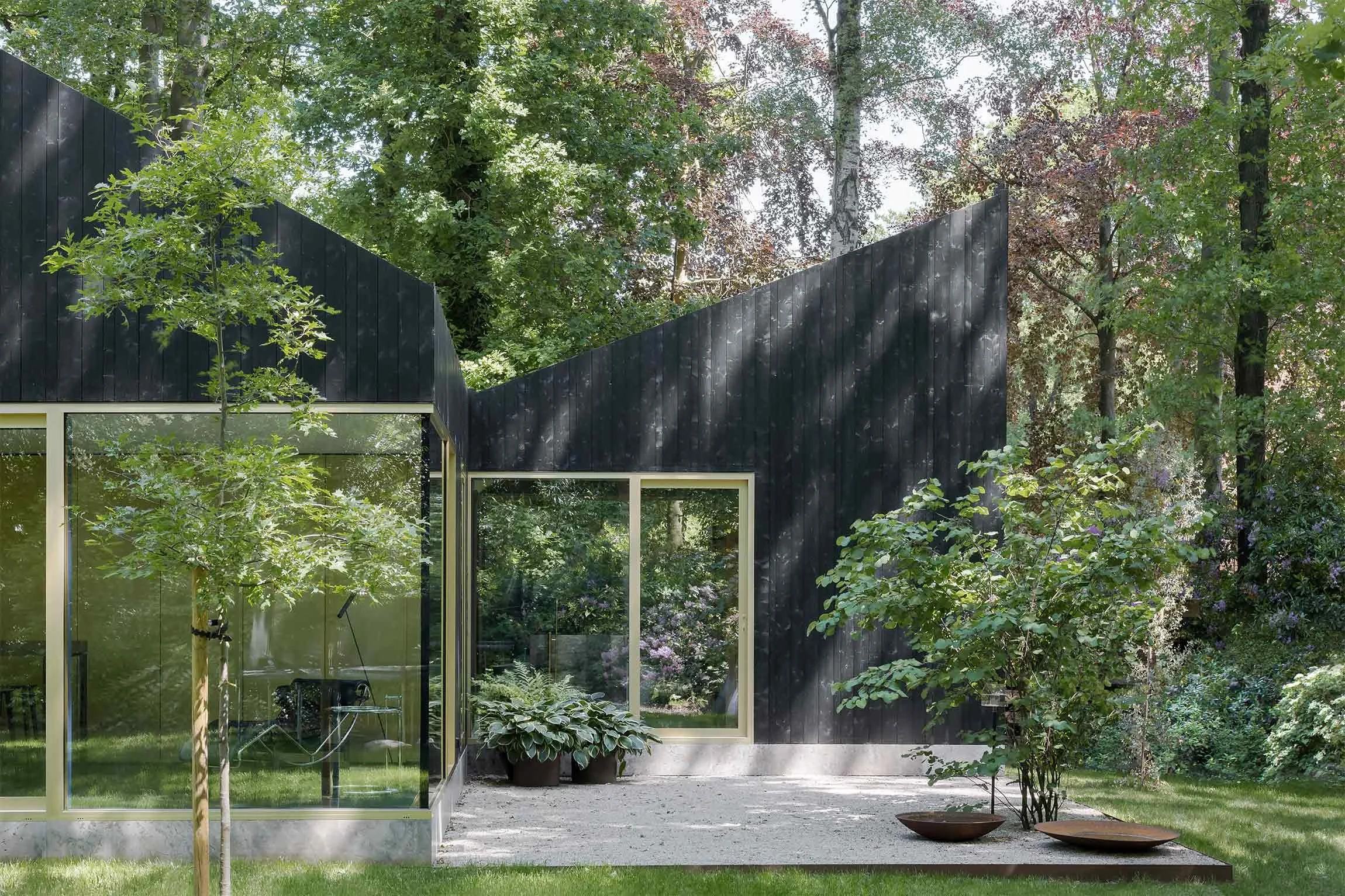 Architecture photo of a renovated chalet with black wooden cladding and large windows surrounded by lush green trees and a luxury garden.