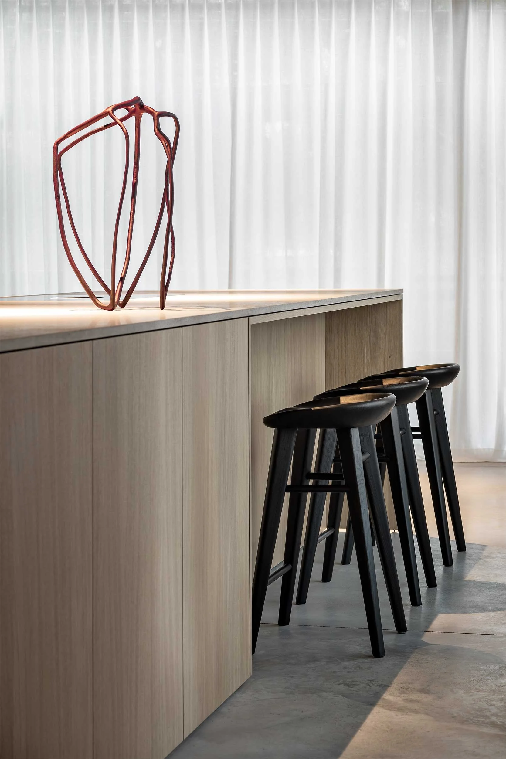 Modern dining or bar area with black bar stools lined up at a light wood counter, decorative art sculpture on the counter, white curtain backdrop, and a concrete floor.